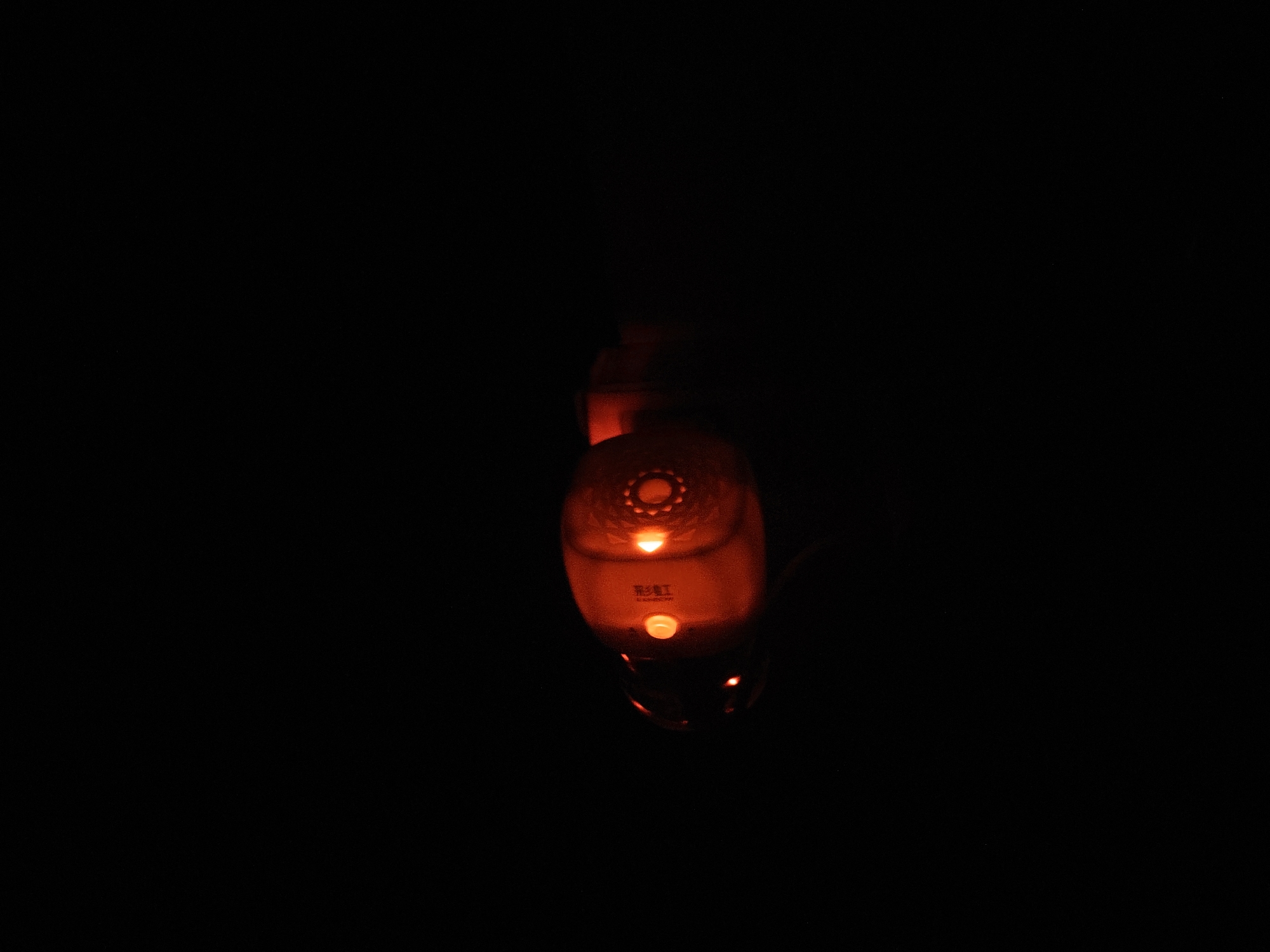 An illuminated object, possibly a lantern or decorative light, hangs in the darkness. The object is predominantly orange and appears to be made of translucent material. It has a rounded shape with some textured details, including what looks like a circular pattern at the top and a small, circular light source in the center. Below this, there are Chinese characters, and another small, illuminated button or light. The object is dimly lit, with its glow casting a faint orange light onto its surface and the surrounding blackness. The lower portion of the object is obscured by shadow and a slight blur, suggesting movement or a shallow depth of field. The scene is extremely dark, with only the object providing any visible illumination.