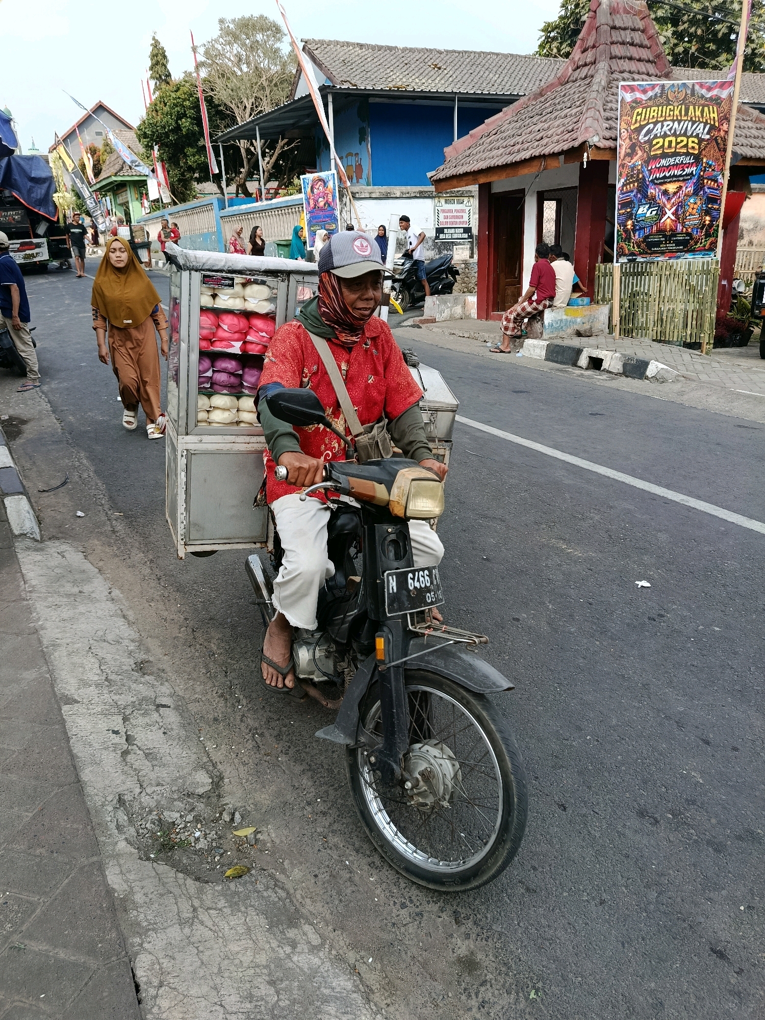 A street scene in Gubukklakah, Indonesia, shows a man on a motorcycle with a cart attached, selling goods. The man, wearing a red shirt, a cap, and a face covering, is riding the motorcycle along a paved road. Attached to the back of the motorcycle is a metal cart filled with stacked items, possibly food or merchandise, appearing to be in shades of pink and white. Beside the road, a woman in a tan hijab and matching outfit walks away from the camera. In the background, there are buildings with tiled roofs, decorative banners, and other people walking or sitting. One banner, prominently displayed on the right side of the image, advertises a "Gubukklakah Carnival 2025 Wonderful Indonesia." The environment appears to be an urban or semi-urban setting with a clear sky.