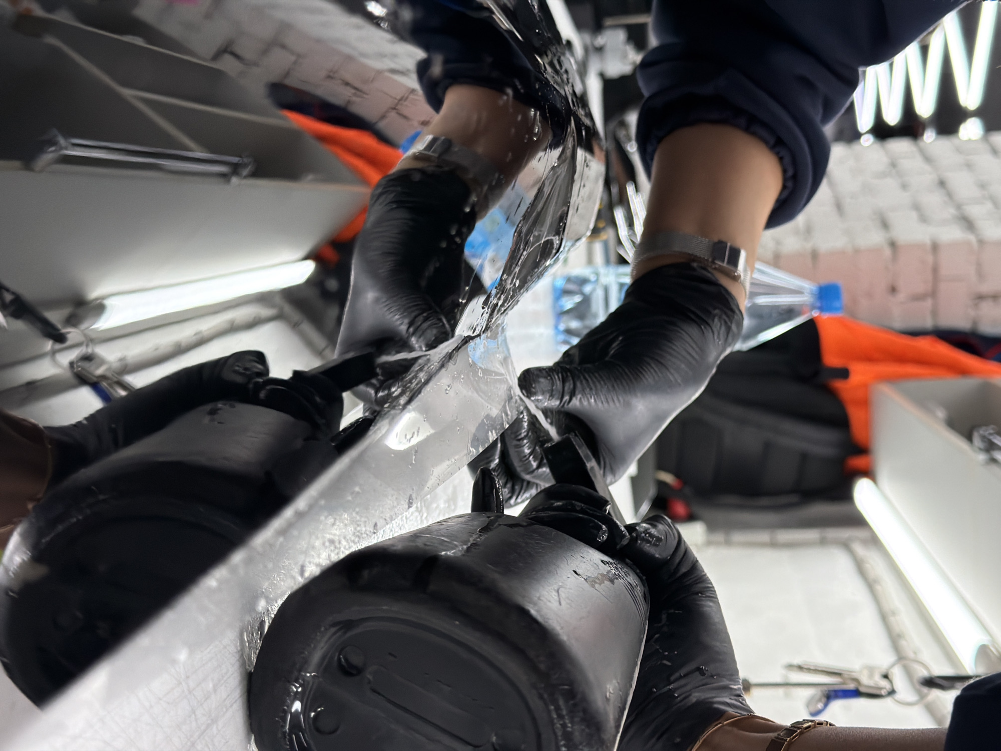 The photograph presents a close-up perspective of two hands, each wearing black latex gloves and a silver wristwatch. They appear to be manipulating a clear, semi-transparent material, possibly plastic film, over a reflective surface. The gloves hold onto dark-colored containers. The setting features fluorescent lighting, a white brick-like structure in the upper right, and components of what seems to be a shelving system on the upper left. Other details include an open water bottle, tools, and a bright orange object. The location is identified as Krasnoyarsk, Russia.