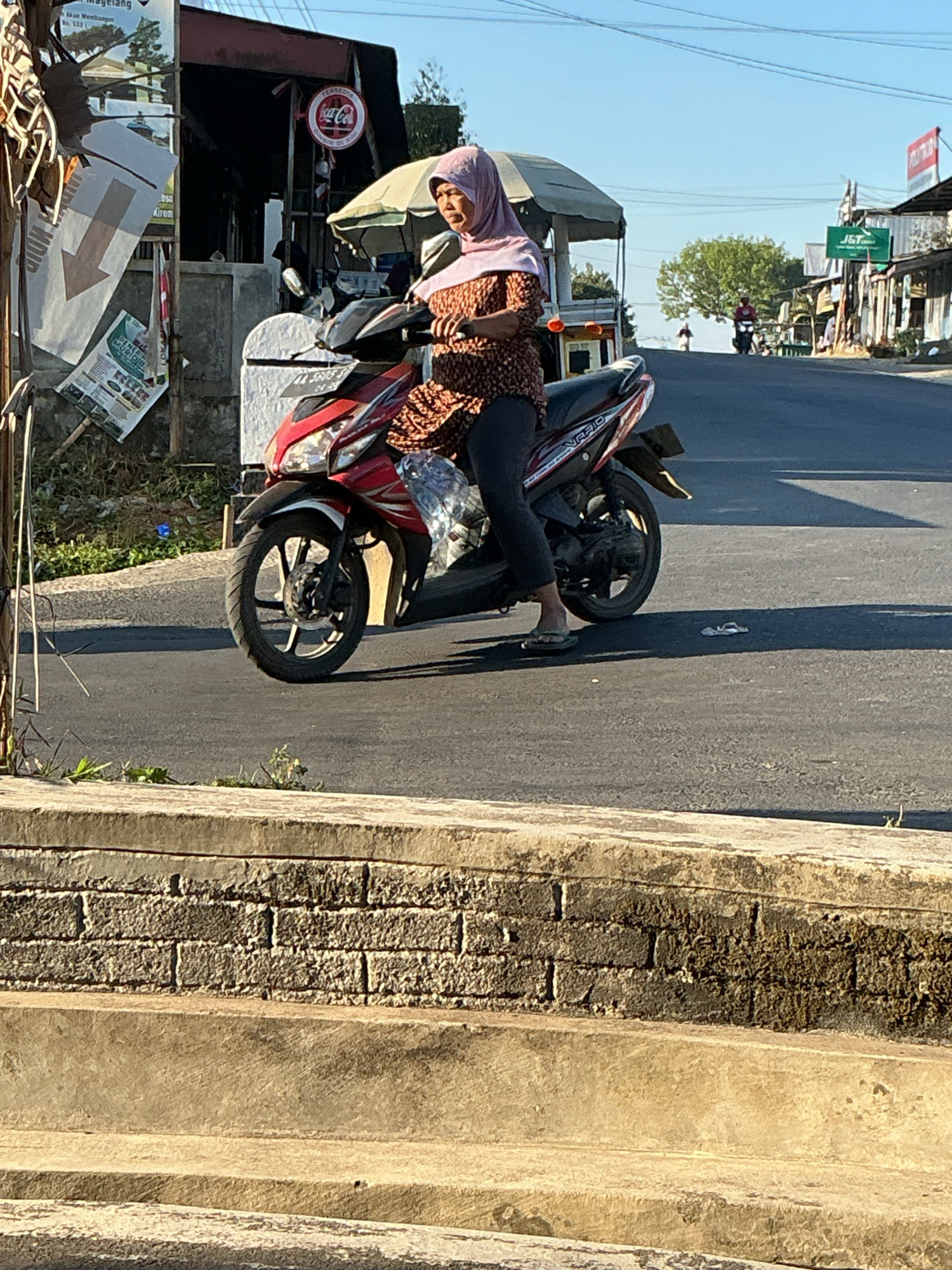 A woman wearing a pink hijab and a patterned long-sleeved shirt is sitting on a red and black motorbike. She is wearing dark pants and flip-flops. She is holding a clear plastic bag filled with something. The motorbike is on a paved road. In the background, there are several buildings, some with signs, and trees. There is also a Coca-Cola sign visible on one of the buildings. The sky is blue. The scene appears to be in a town or city in Kebonrejo, Indonesia.