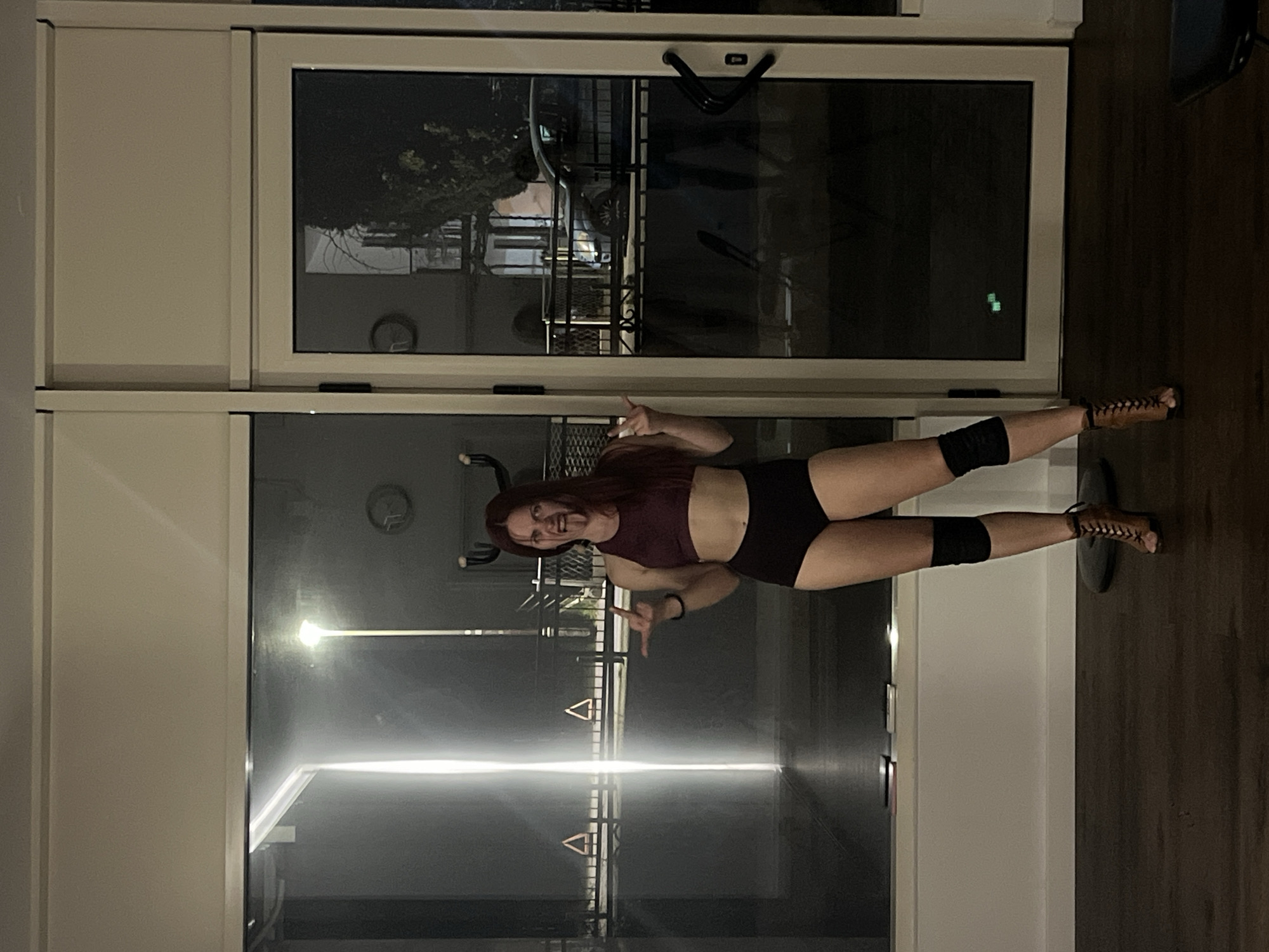 A woman stands in a dance studio in Budva, Montenegro. The studio features large, floor-to-ceiling windows, which provide a view of the city at night. Outside, a parked car is visible behind a fence. The studio is illuminated by interior lighting, creating a bright, reflective surface on the windows. The woman wears a crop top, shorts, knee pads, and laced high heels, and poses with both hands up. The flooring appears to be dark wood or laminate.