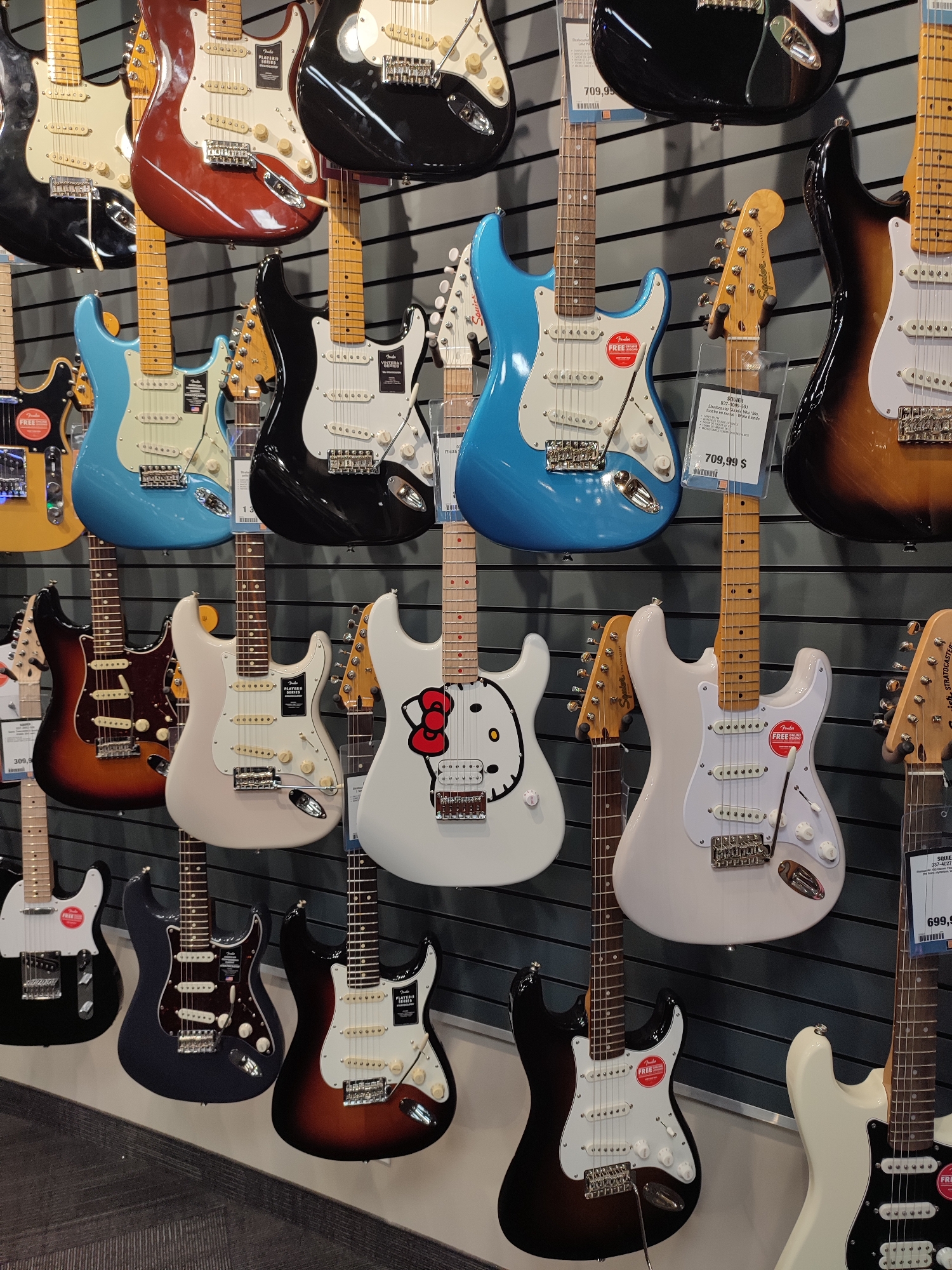 The image shows a wall display of electric guitars in a store, likely a musical instrument shop. The guitars are of various colors, including black, red, blue, white, and sunburst. They are all electric guitars, most of them appearing to be Fender Stratocasters or similar models, hung on a grey slatwall. Some guitars have price tags or information cards attached to them. The lighting suggests an indoor environment, with the guitars illuminated from the front. The overall presentation is neat and organized, typical of a retail display. The scene does not contain any people or visible actions. The location is specified as Longueuil, Canada.