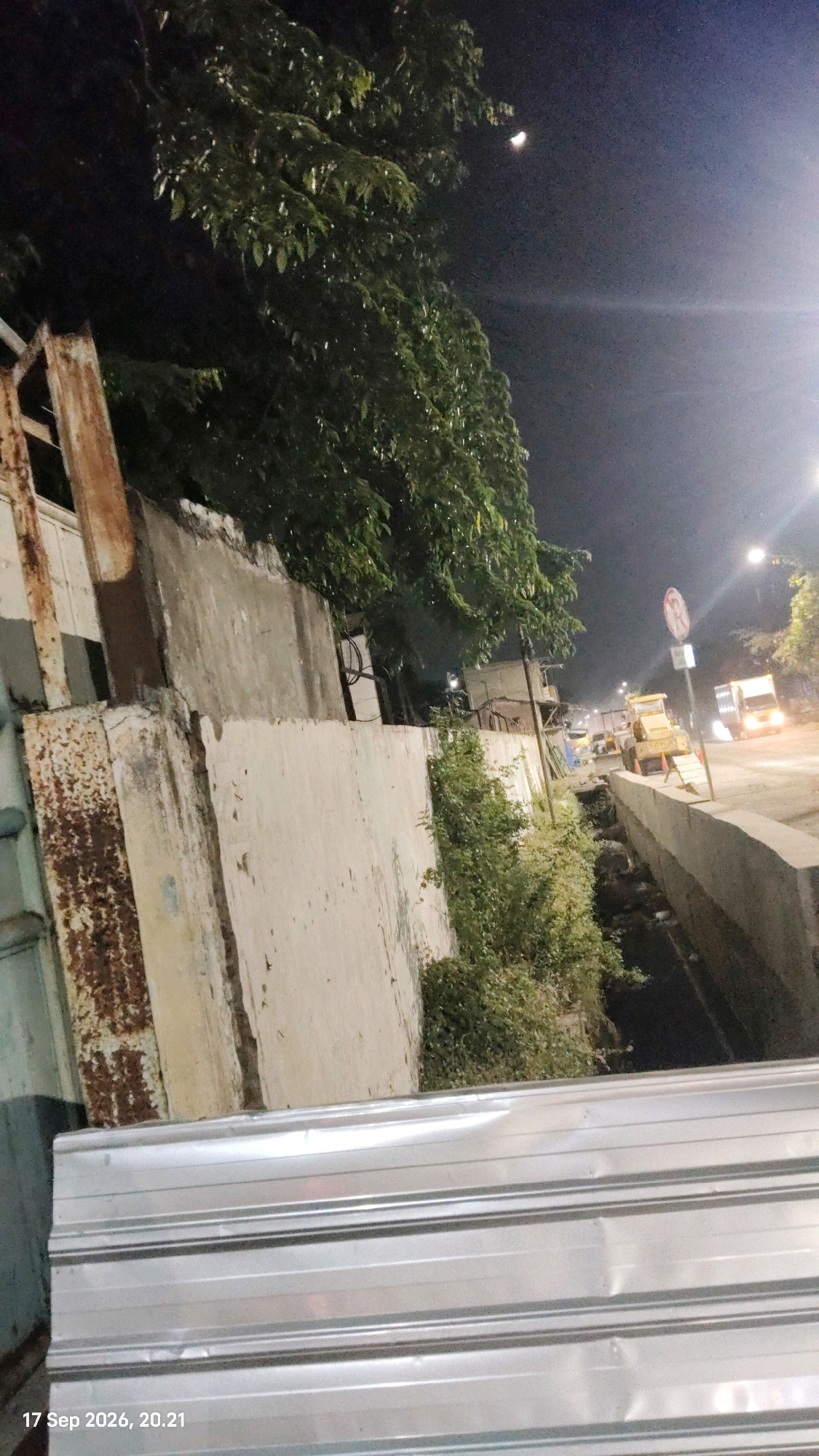A nighttime street scene in Surabaya, Indonesia, depicts a construction site alongside a road. In the foreground, a corrugated metal barrier is prominent. Behind it, a weathered concrete wall, showing signs of rust and damage, separates the viewer from a narrow, debris-filled drainage ditch. Overgrown green vegetation grows along the ditch and the wall. The background reveals a street with active construction machinery, including a yellow bulldozer. Vehicles, including a truck and a yellow transport vehicle, are visible on the road, illuminated by streetlights and headlights. A faint crescent moon is visible in the dark, hazy sky, along with a traffic sign with a red circle and a diagonal line, indicating a prohibition. The timestamp "17 Sep 2026, 20.21" is present in the lower left corner.