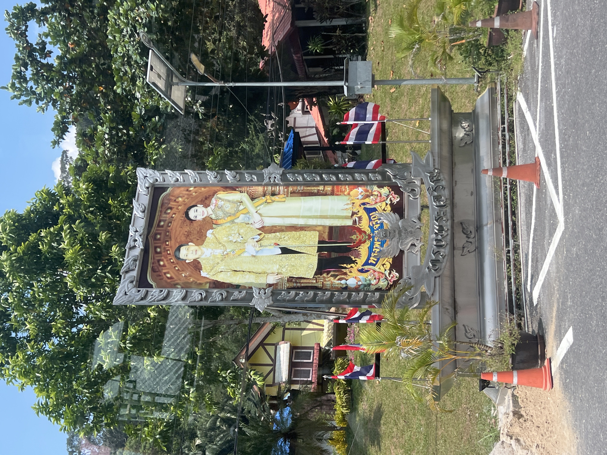 A large, ornate framed portrait of a man and a woman in traditional Thai attire is displayed outdoors in Tambon Wang Prachan, Thailand. The man, on the left, wears a golden robe with intricate patterns and a sash, while the woman, on the right, wears a light-colored traditional dress and a sash. Both figures are looking forward with neutral expressions. The portrait is mounted on a multi-tiered silver pedestal adorned with decorative elements.
In the background, lush green trees and foliage dominate the scene, with a clear blue sky overhead. Behind the portrait, a small, yellow and brown building with a red roof is visible. Several Thai flags are planted in the ground in front of the portrait. A modern solar-powered street light stands to the right of the portrait, and there is a gray electrical box attached to its pole.
In the foreground, the scene transitions to a paved road with white lane markings. Two orange traffic cones are placed on the road, one near a potted plant and the other further to the right. The ground around the traffic cones is covered with sand and gravel. The overall environment appears to be a public space, possibly a roadside attraction or a monument display area.
