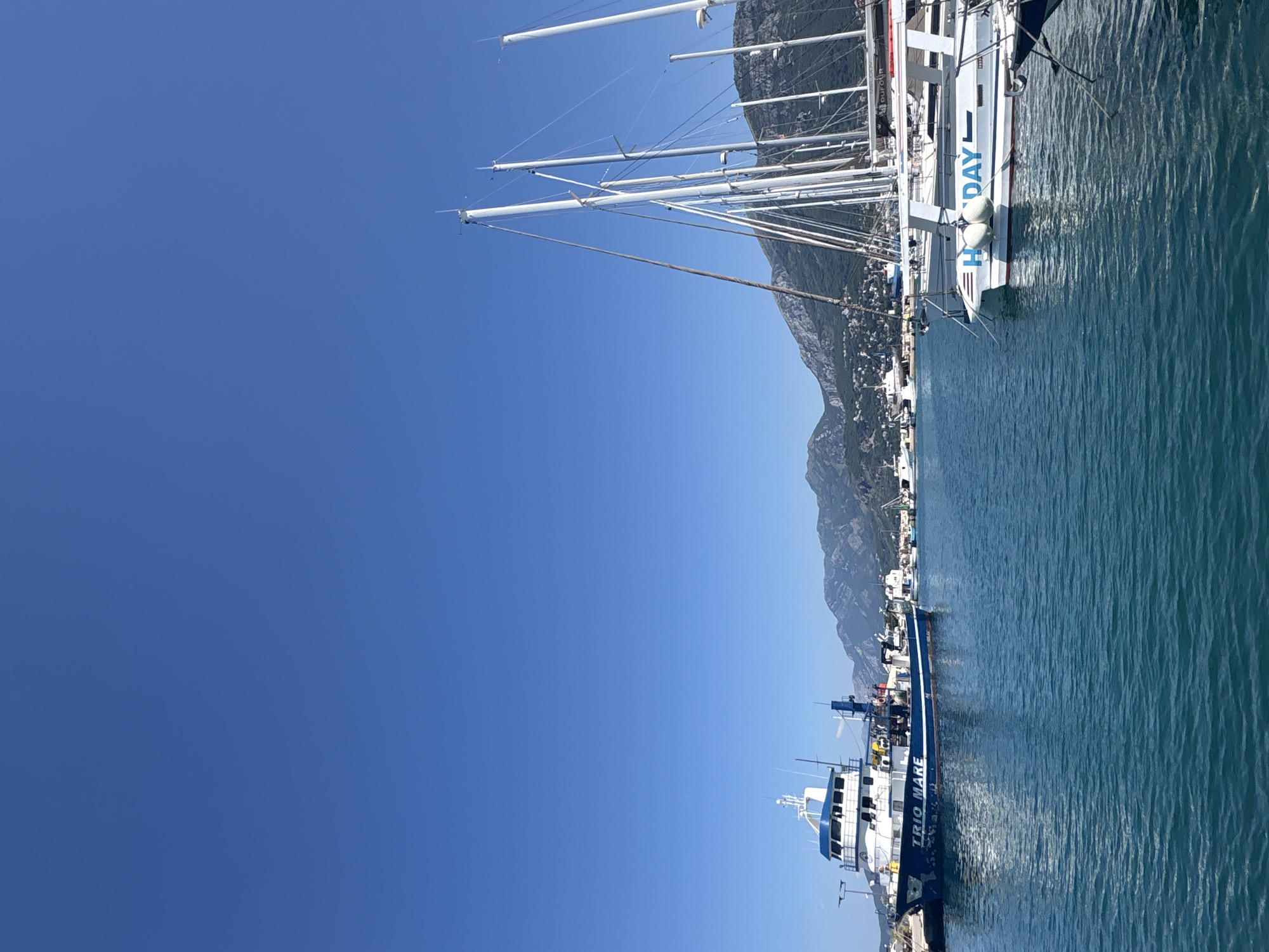 The image shows a harbor in Bar, Montenegro, on a clear, sunny day. The sky is a vibrant blue, and the water is a deep turquoise. Several boats are docked in the harbor, including a large blue and white fishing trawler named "TRIO MARE," and several white sailboats with tall masts. In the background, a lush green mountainside rises behind the town, which is dotted with buildings. The scene is peaceful and serene, with no people visible in the frame. The boats appear to be well-maintained and ready for use. The overall impression is one of a tranquil coastal town on a beautiful day.