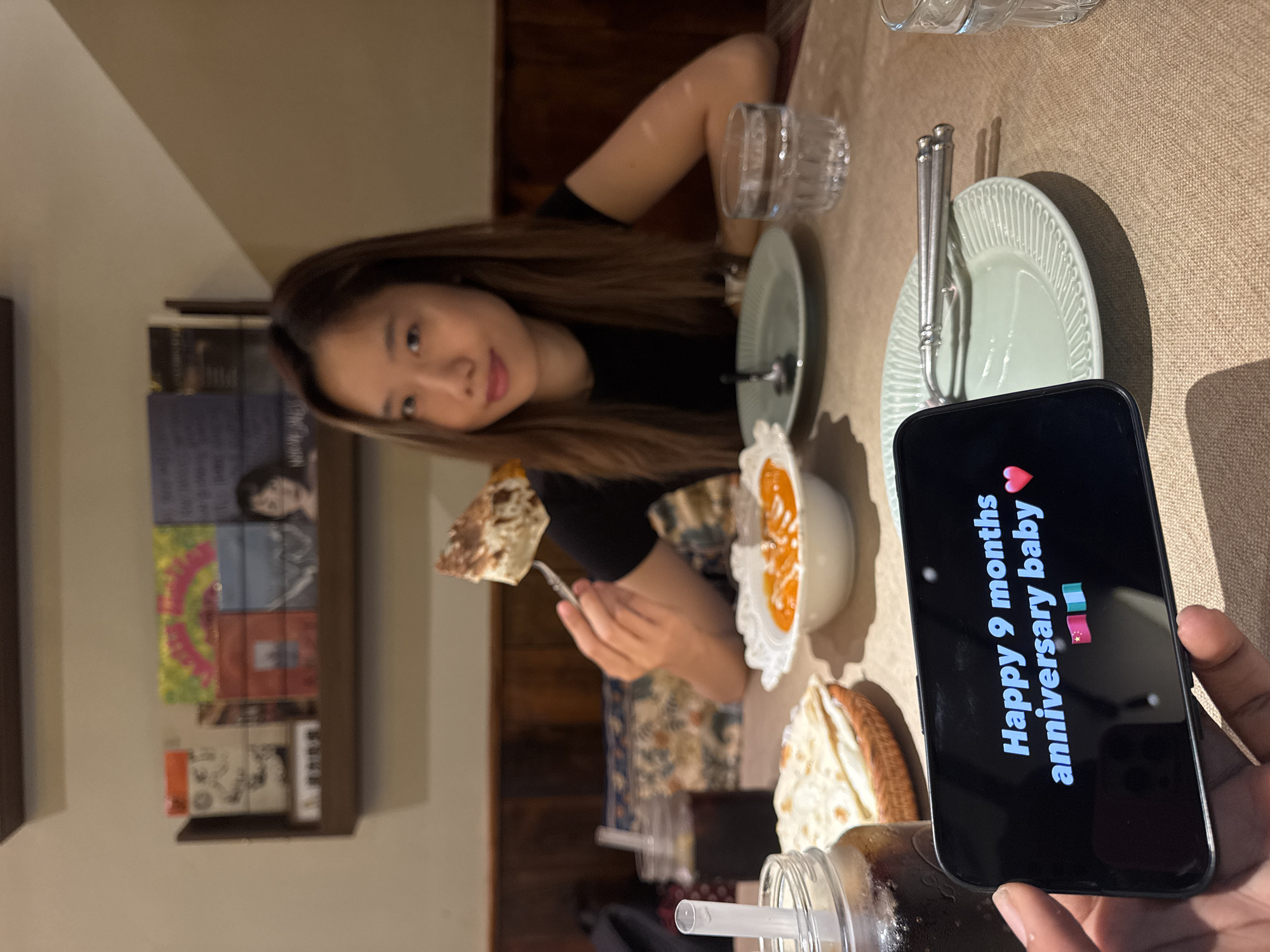 A woman sits at a table in what appears to be a restaurant in the Yuzhong District, China, eating food with a fork. The table is set with plates, silverware, and drinks. A mobile phone with the phrase "Happy 9 months anniversary baby" with heart and flag emojis in Chinese is in the lower right. In the background is a bookshelf, and wood walls. The lighting is warm and dimly lit.