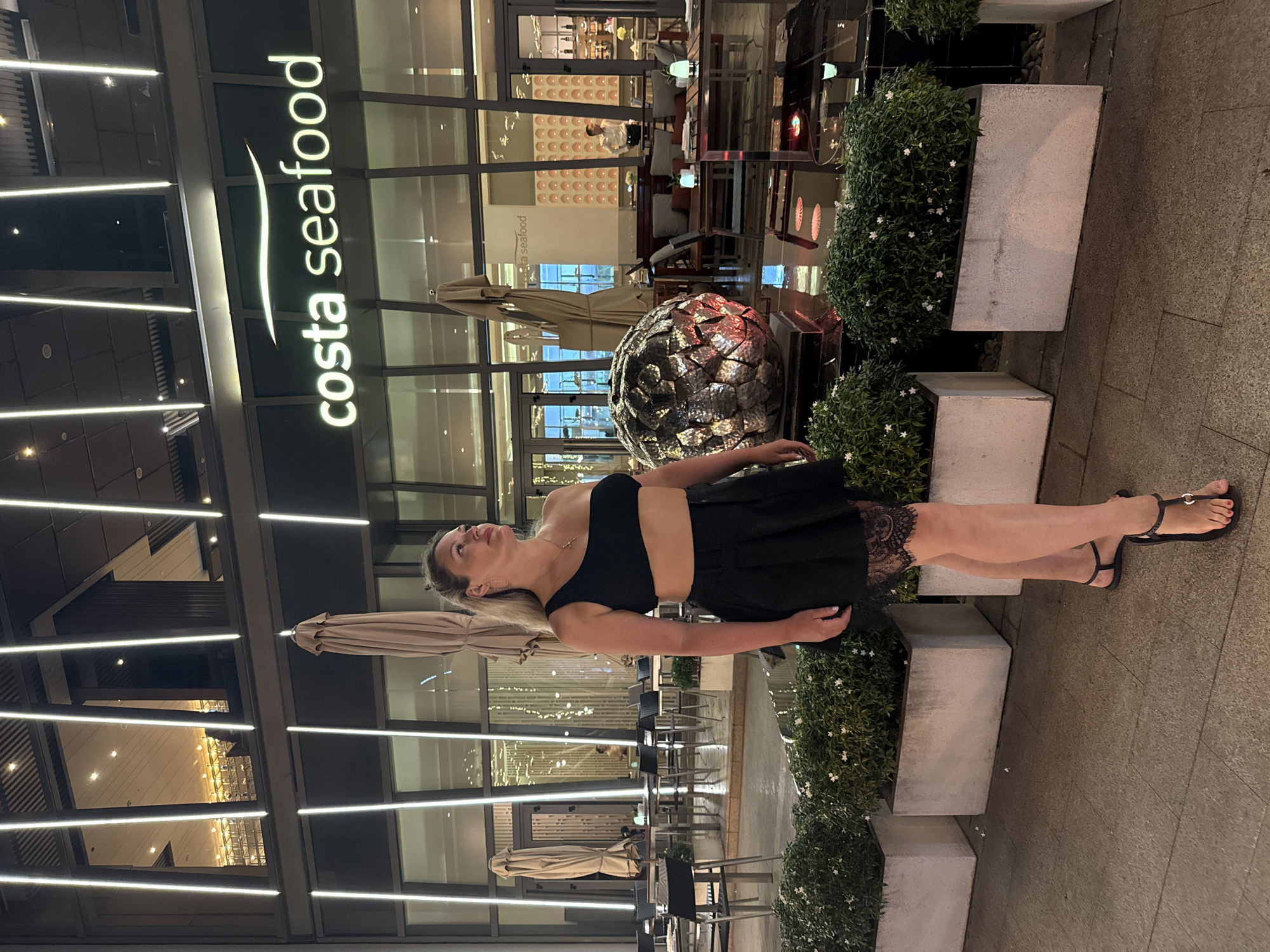 A woman in a black outfit is standing on a sidewalk outside Costa Seafood in Nha Trang Ward, Vietnam. The restaurant's signage is visible, as are outdoor dining areas with umbrellas and furniture. Potted plants line the sidewalk. The woman is wearing black sandals.