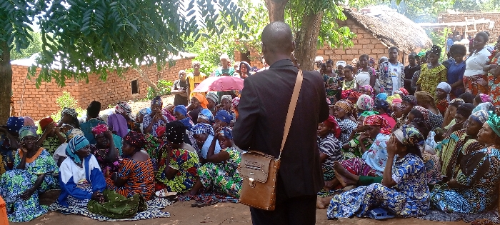 A crowd of women, many in colorful patterned clothing and head coverings, sit on the ground. A man in a dark suit faces them, his back to the viewer, with a satchel slung over his shoulder. They gather in a clearing surrounded by trees, with simple brick buildings and thatched roofs visible in the background. An umbrella is visible in the background, and the sun shines down on the assembled group.