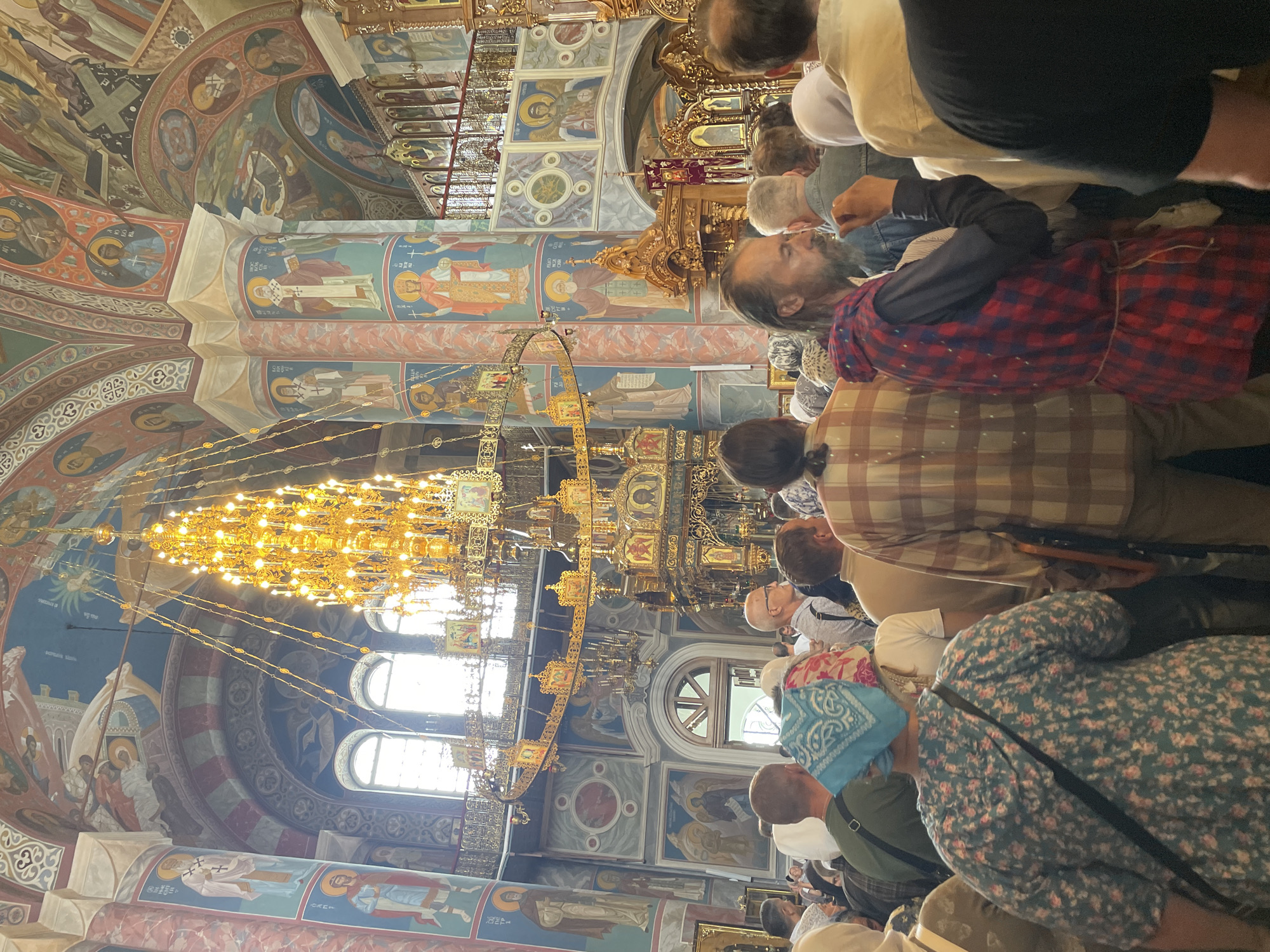 The image depicts the interior of the Holy Trinity Cathedral in Diveyevo, Russia, during a gathering. A large, ornate golden chandelier hangs prominently from the ceiling, surrounded by elaborate religious murals and architectural features. A crowd of people, mostly women, are gathered inside the cathedral, facing towards the front. Many are wearing headscarves, typical for attending religious services in Orthodox churches. The atmosphere appears to be one of reverence and participation in a spiritual event.