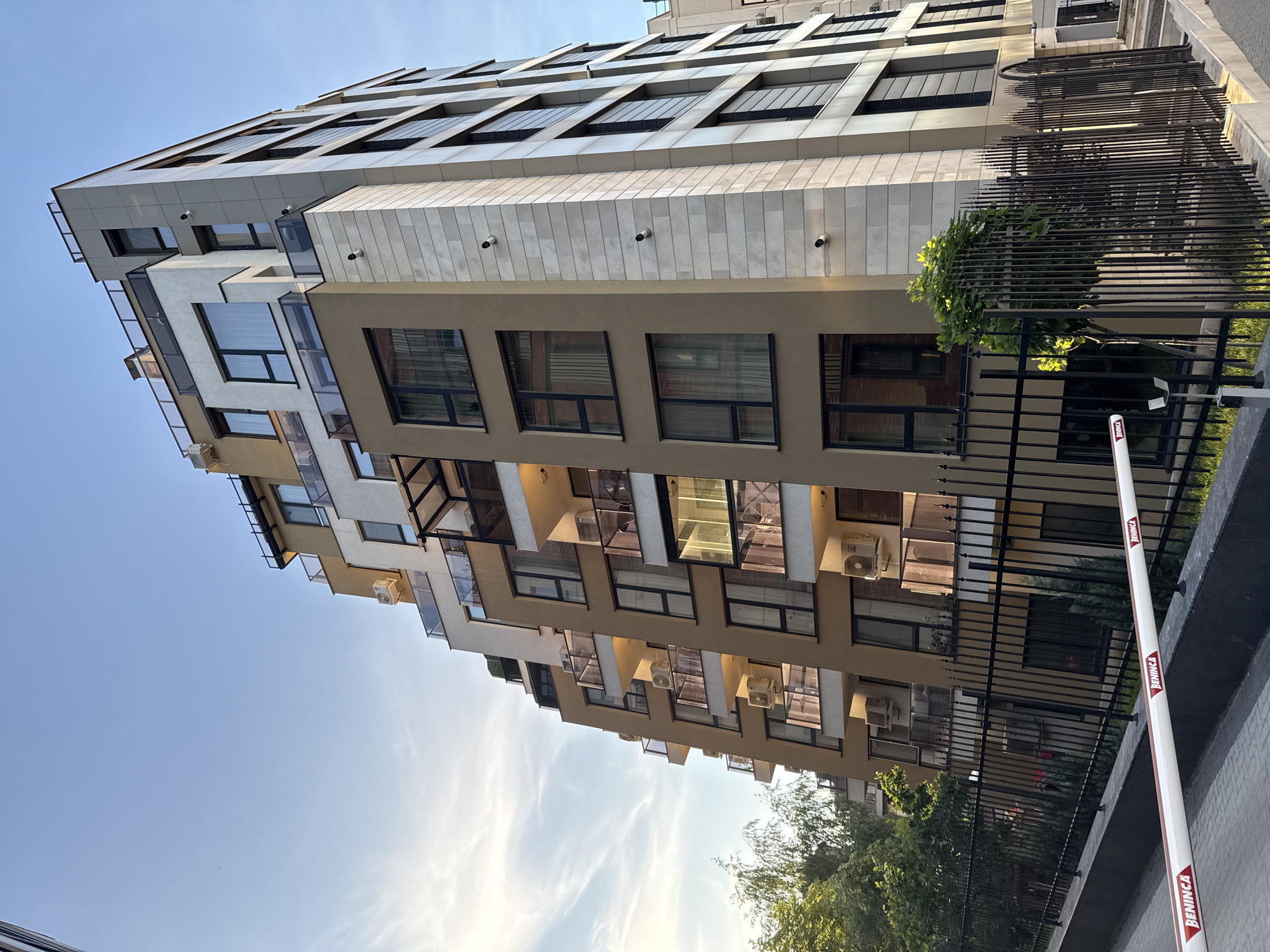 The image depicts a modern apartment building in Sofia, Bulgaria. The building has a multi-tiered design with a mix of neutral-colored facades, including beige, white, and gray panels. It features numerous windows and balconies with transparent railings. Air conditioning units are visible on the exterior walls of some apartments.
A black metal fence runs along the perimeter of the property, with a white and red automatic barrier gate in the foreground. Lush green trees and vegetation are visible behind the fence, adding a touch of nature to the urban setting. The sky is clear with a few wispy clouds, suggesting a sunny day. No people are visible in the frame.