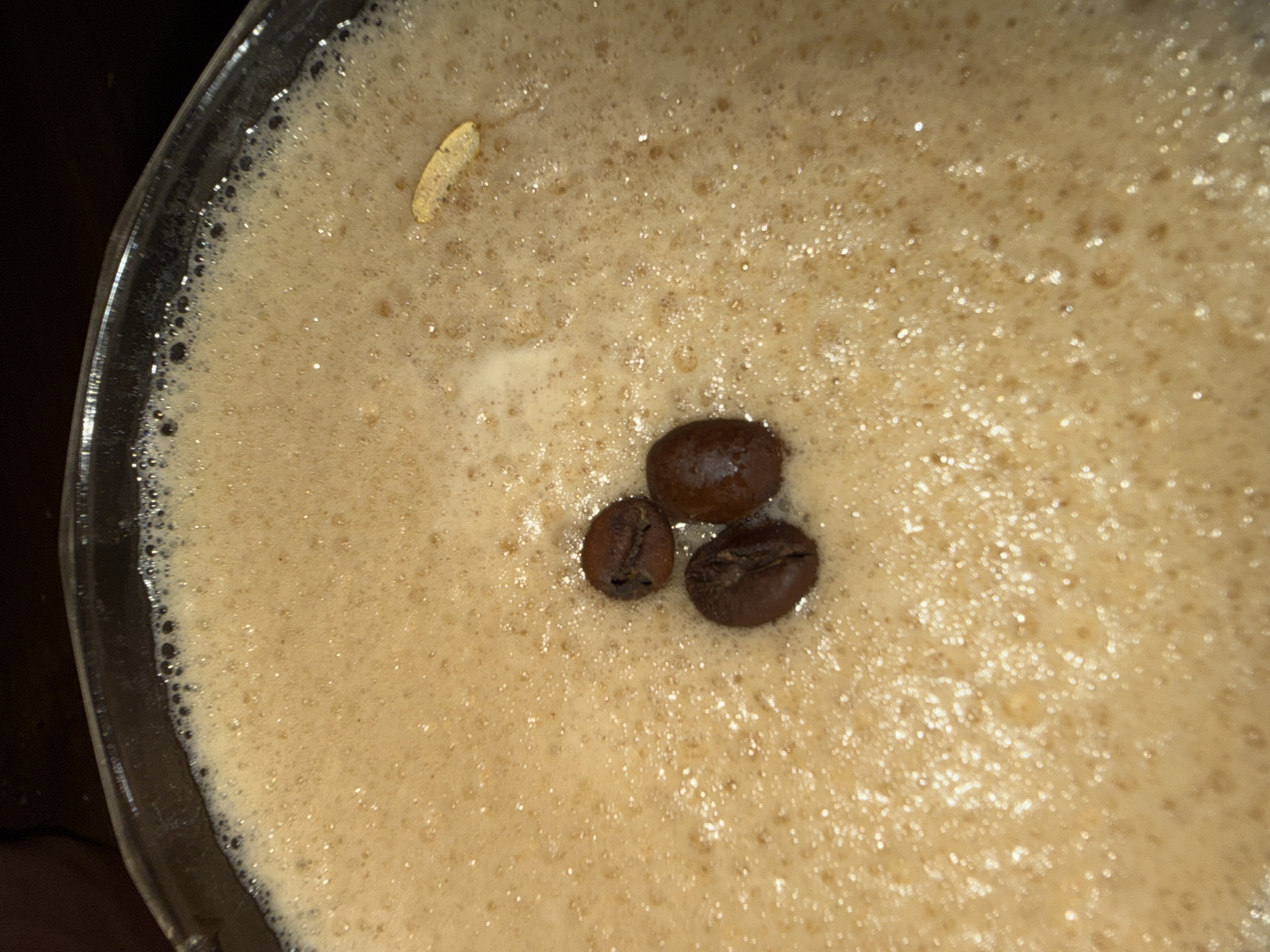 A close-up, top-down shot captures the foamy surface of a beverage in a clear glass. The drink features a thick layer of light brown froth, densely packed with small bubbles. Three dark brown, whole coffee beans are clustered near the center of the foam, appearing glossy and slightly wet. In the upper left quadrant of the foam, a single, elongated, yellowish object, possibly a spice like a cardamom pod or a piece of lemon zest, floats on the surface.
The rim of the clear glass is visible along the left and top-left edges of the frame, showing the contrast between the light-colored foam and the dark, out-of-focus background. The lighting is somewhat dim, creating highlights on the coffee beans and a sparkling effect on some of the foam bubbles. No people or text are visible in the image. The scene suggests a prepared drink, likely a coffee-based beverage or a cocktail, presented with a garnish.
