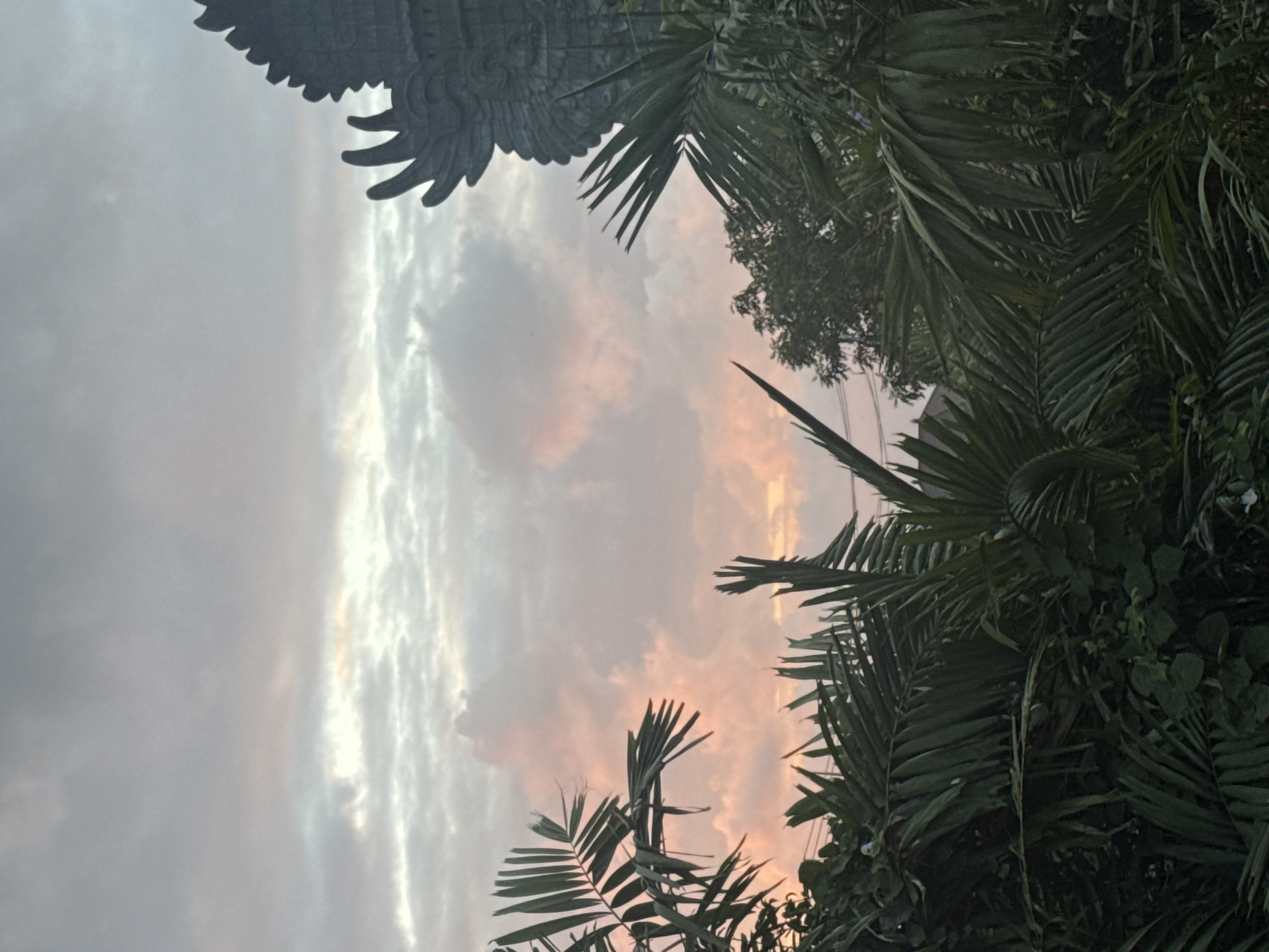 The image shows a view of the sky at sunset through lush tropical foliage. In the upper right corner of the frame, a portion of a large stone sculpture, likely a Garuda statue, is visible. The sculpture is dark gray and detailed, with a winged headdress. The sky is filled with soft, pastel-colored clouds, primarily in shades of pink, orange, and gray, indicating the approaching twilight. Dense green palm fronds and other tropical leaves fill the foreground and frame the sky, creating a sense of depth. The overall atmosphere is serene and tropical. This scene is consistent with views found in Kuta Selatan, Indonesia, known for its beaches and the iconic Garuda Wisnu Kencana Cultural Park, which features a massive Garuda statue.