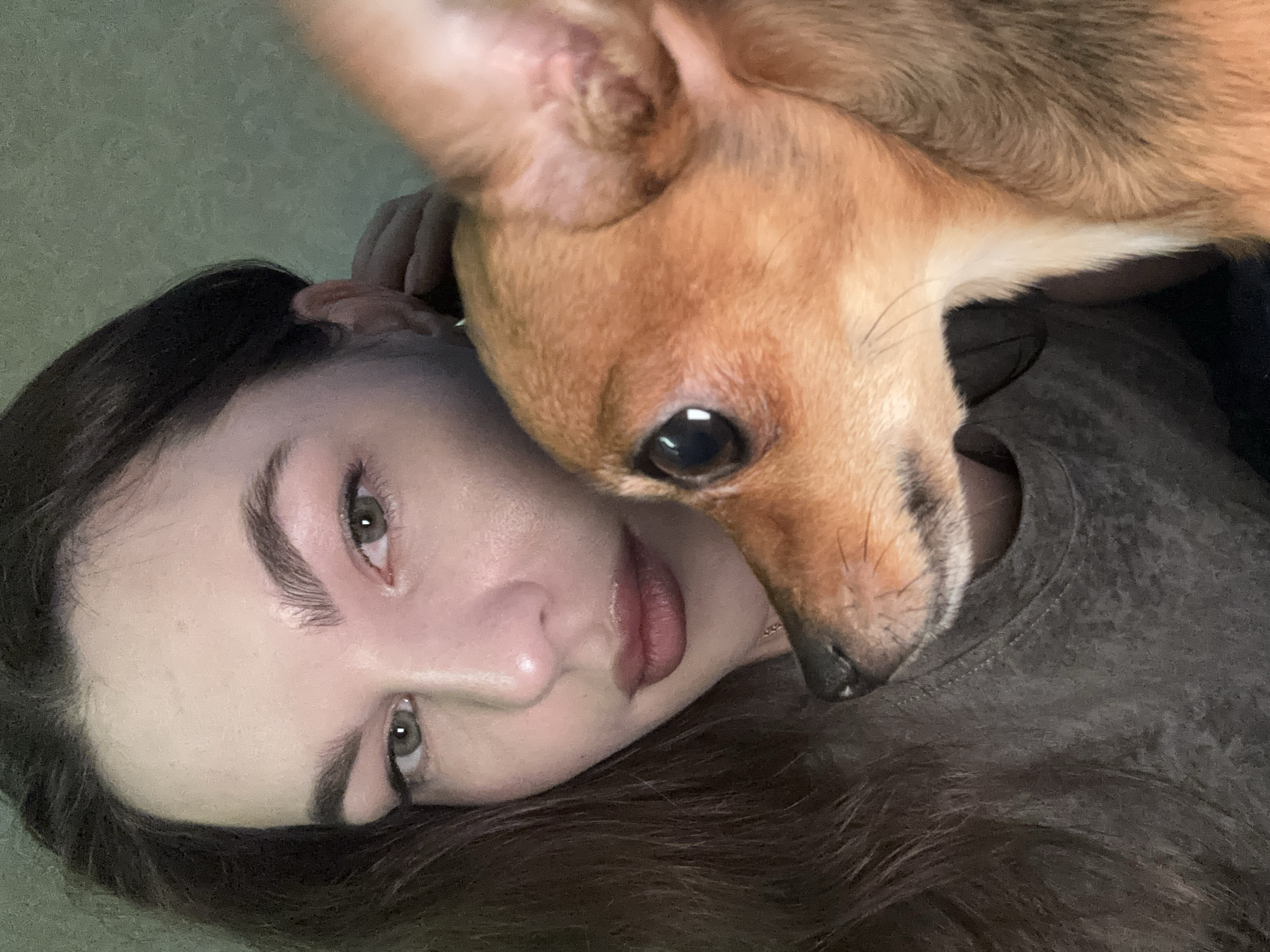 A young woman with dark hair and olive-toned skin lies with her face tilted towards the camera. A small, light brown dog rests its head and snout on her chest and shoulder. The woman wears a gray t-shirt. The background appears to be a textured, muted green wall. There is no evidence to support the location as Chaikovsky, Russia.