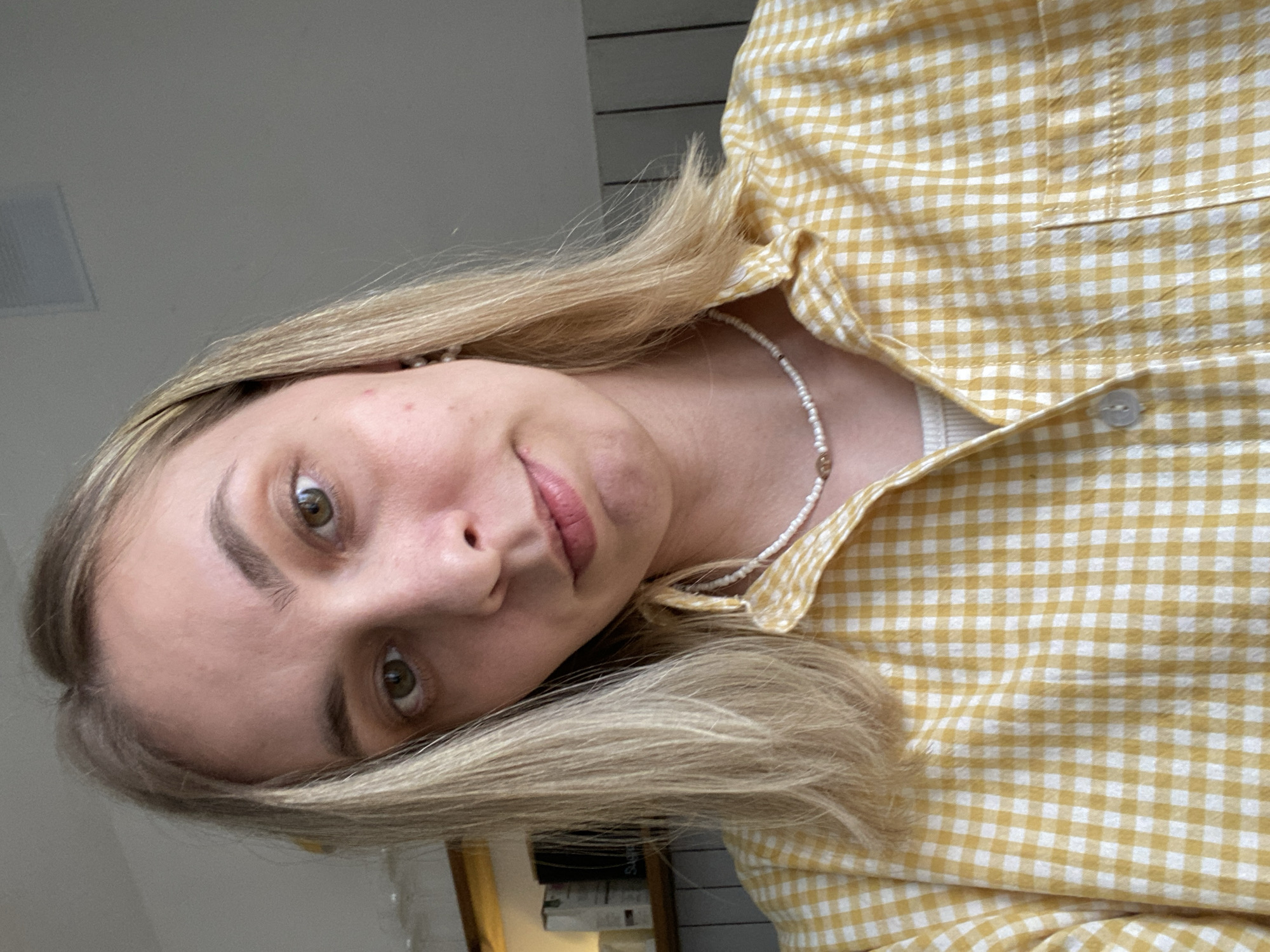 A woman with light blonde hair is facing the camera, wearing a yellow and white checkered shirt and a beaded necklace. The background is a light, neutral color. The image is taken indoors, possibly in a home. There's a glimpse of a white square, perhaps an air vent or lighting fixture, in the upper left. Part of a bookcase with books is visible in the lower left. There is no visible evidence of a location in the city of Chaikovsky, Russia.