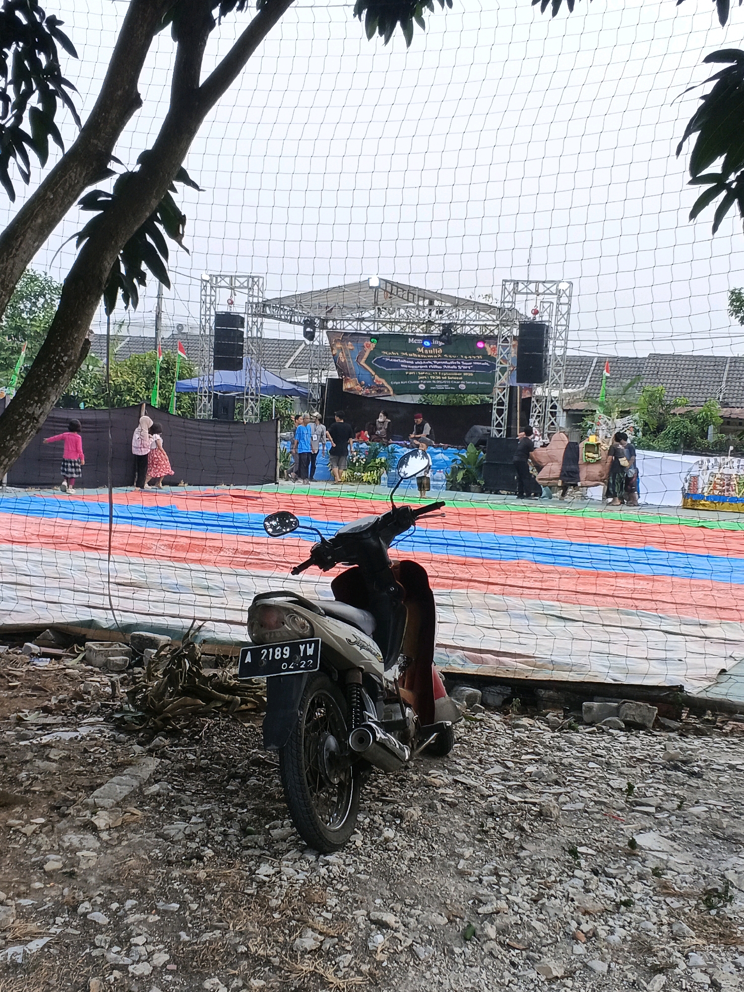 The image captures an outdoor scene at what appears to be a community event in Panamping, Indonesia. In the foreground, a parked motorcycle with license plate "A 2189 YW" and an expiration date of "04-22" is visible on a gravelly ground. Behind the motorcycle, a stage setup is present, with a large banner displaying text, possibly related to a religious or cultural celebration. Several individuals are seen on or around the stage, some appearing to be performers or staff. Colorful fabric, likely part of decorations or ground cover, is spread out in front of the stage. In the background, trees and buildings suggest a residential or semi-urban environment. The overall lighting indicates an overcast day.