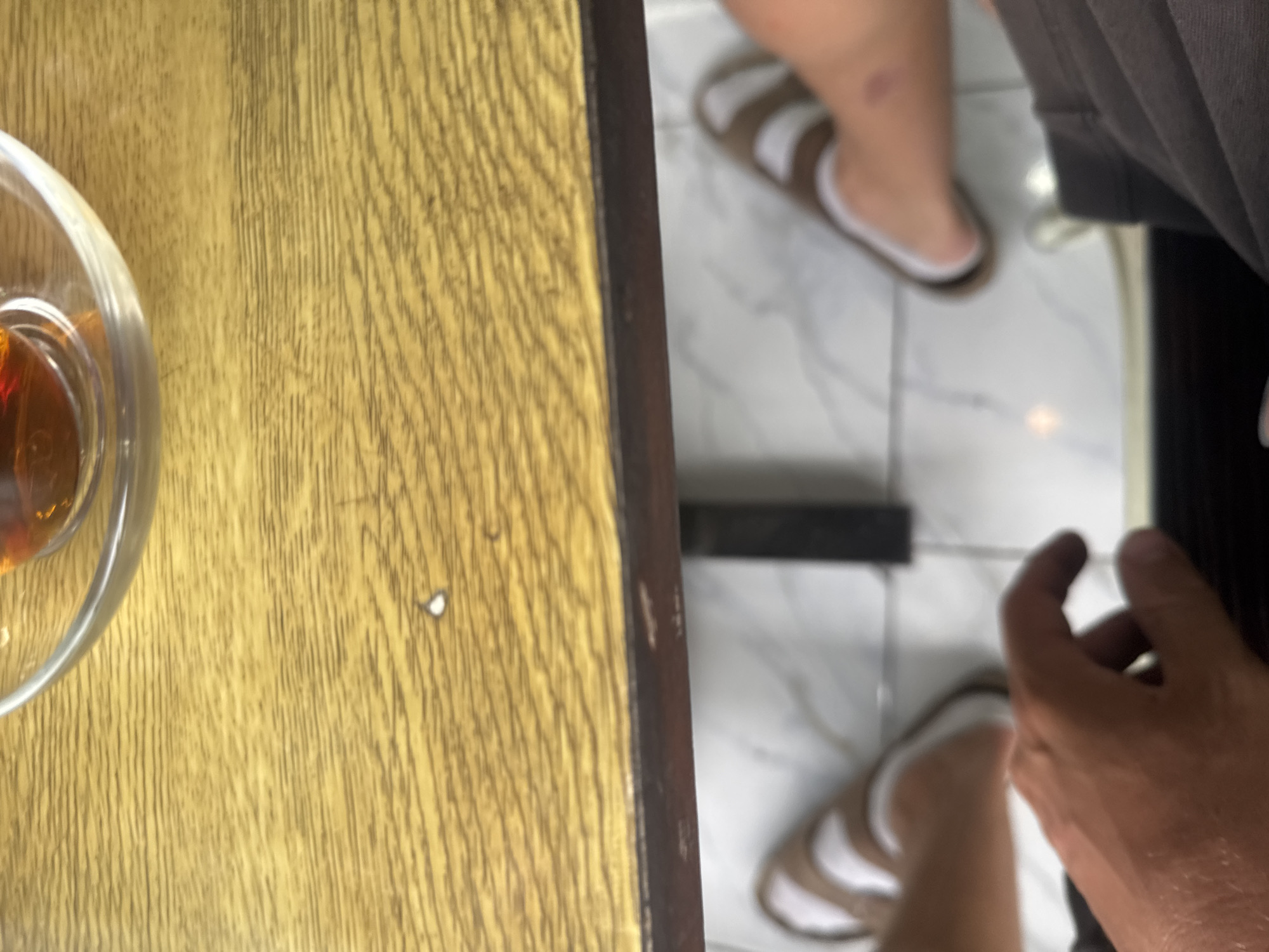 The image captures a close-up of a person's hand resting on a wooden table, with a glass of amber liquid on the left side of the frame. In the background, another person's legs and feet are visible, wearing white and brown striped sandals. They are standing on a tiled floor. The scene appears to be indoors, possibly in a casual setting like a cafe or restaurant. The lighting suggests it could be daytime. The focus is on the textures of the wood and the tiled floor, as well as the hands and feet. There are no visible signs or text in the image. The overall mood seems relaxed and informal.