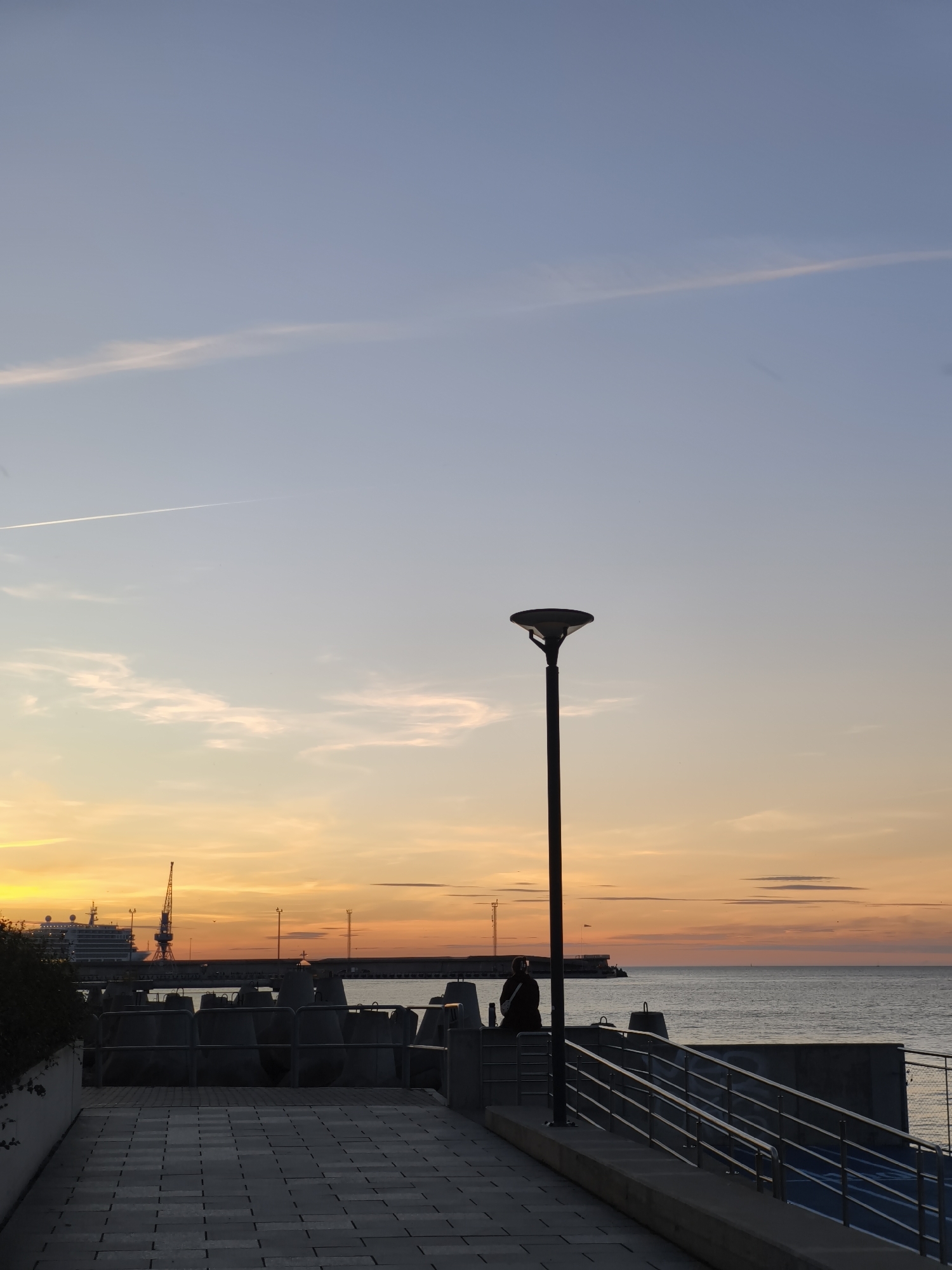 The image depicts a sunset over the Baltic Sea in Tallinn, Estonia. In the foreground, a paved walkway leads towards the sea, bordered by a metal railing. A lone street lamp stands to the right, silhouetted against the vibrant orange and yellow hues of the sunset. A person is seated on a wall, facing the sea, with their back to the camera. In the middle ground, a breakwater with concrete tetrapods lines the coast, and a large cruise ship is docked in the distance, with a crane beside it. The sky is a gradient of blue and orange, with a faint contrail visible high above. The sea is calm, reflecting the colors of the sunset.