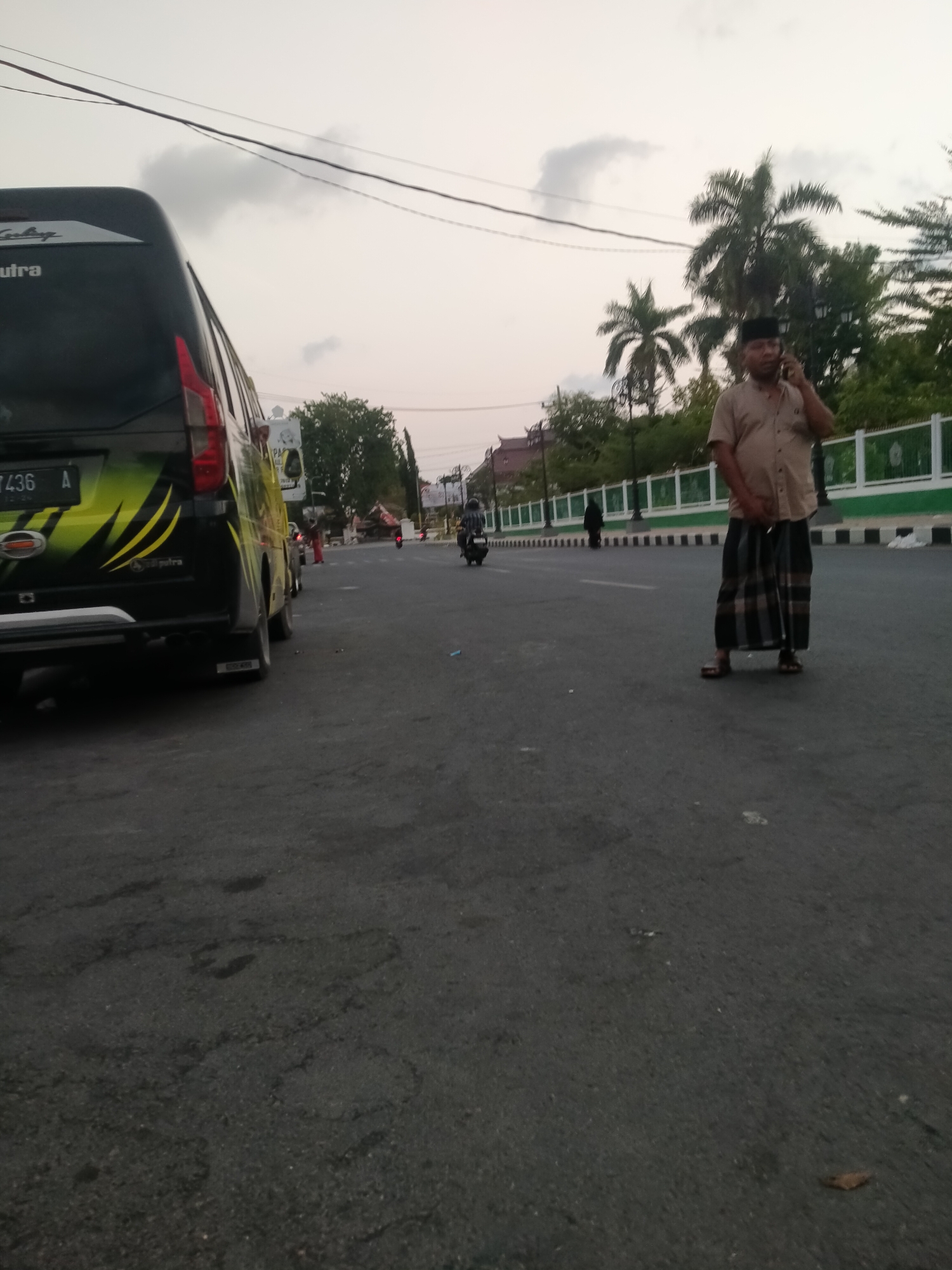 The image depicts a street scene in Pamekasan, Indonesia, during the late afternoon or early evening, as indicated by the diffused lighting. In the foreground, the rear of a black van with yellow and green decals is parked on the right side of the asphalt road. The van has a license plate that reads "7436 A". To the right of the van, a man wearing a light brown short-sleeved shirt, a black peci (cap), and a sarong with a checkered pattern is standing and speaking on a mobile phone. He is facing towards the right of the frame and is wearing brown sandals. In the background, there is a white and green fence lining the street, with trees and some buildings visible beyond it. Further down the road, a person on a motorcycle is moving away from the camera. The sky is overcast with some diffused clouds. The overall environment appears to be an urban setting with a road, vehicles, pedestrian traffic, and some greenery.