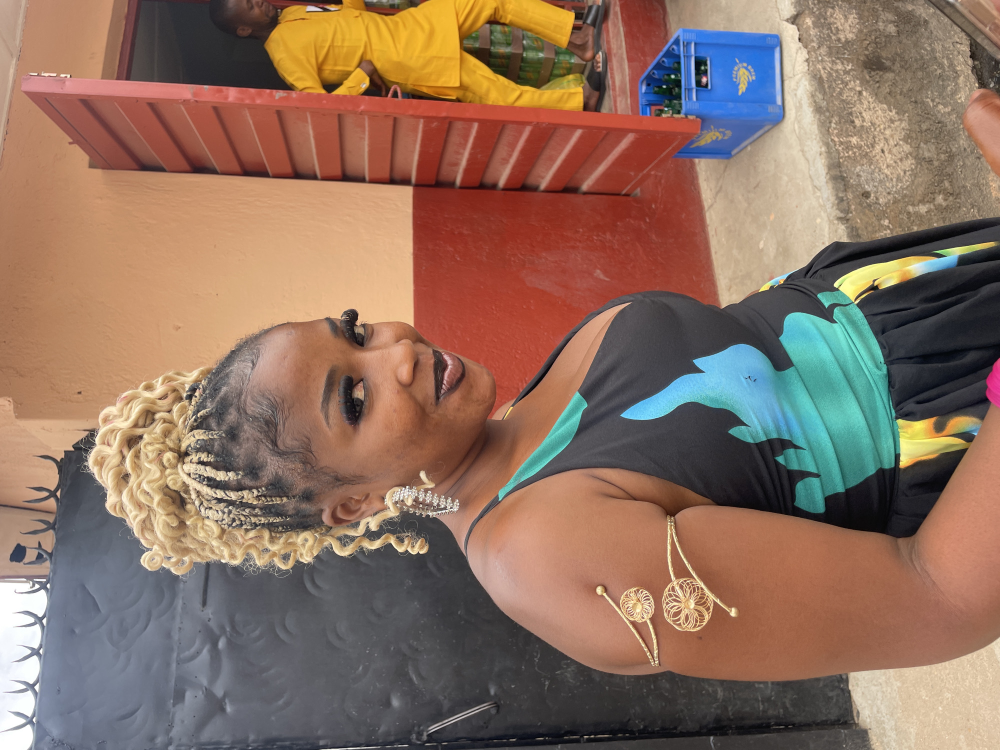 A woman with blonde braided hair, wearing a black dress with blue and green patterns, and gold arm jewelry, is the central subject. A man in a yellow suit lies horizontally in an open window above her. To the side of the window is a blue crate filled with green bottles. The scene appears to be outdoors, possibly in Jos, Nigeria, given the context. The woman is looking towards the camera. A black metal gate is visible near her. The building behind them is painted in sections of orange, red, and peach.