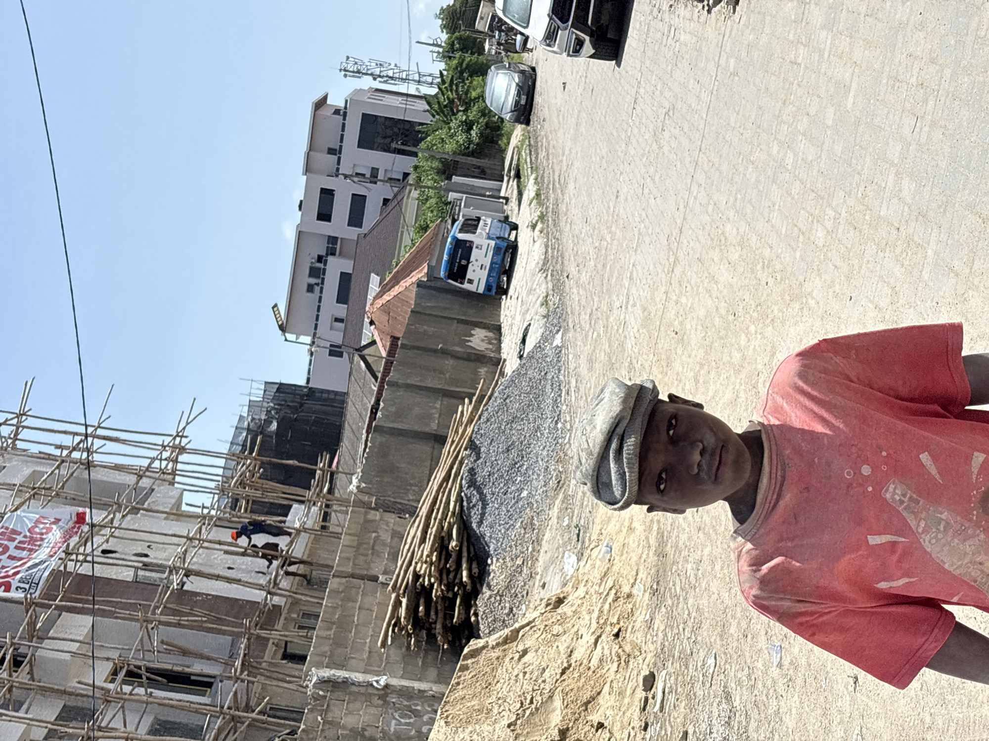 The image shows a young person wearing a red t-shirt and cap, standing near a construction site in Igboefon, Nigeria. The site includes scaffolding and unfinished concrete structures. There are piles of building materials, including gravel and wooden poles, alongside a paved road with parked vehicles. The sky is clear.