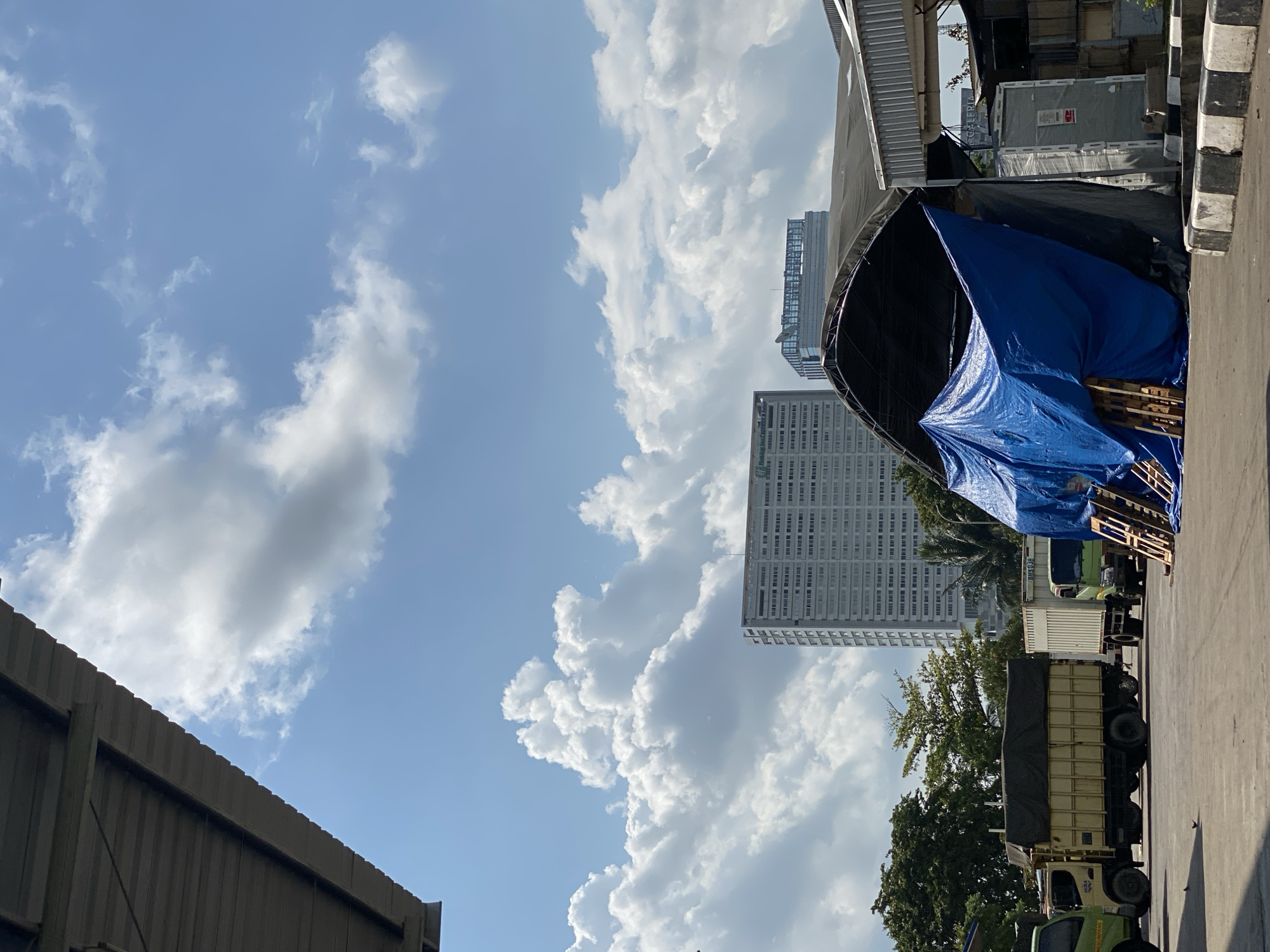 This image depicts an outdoor scene likely in the vicinity of Gambut Barat, Indonesia, on a clear day with scattered clouds. In the foreground, a large, industrial-style awning extends from the left side of the frame, casting a shadow. Beneath this awning, a prominent blue tarp covers a structure, with wooden pallets stacked nearby. To the left of the tarp, two trucks are parked on a paved surface; one is a yellow truck with a covered cargo bed, and the other is a smaller white truck. Further in the background, a tall, modern white building with numerous windows dominates the skyline, and another glass-fronted building is visible behind it. Green trees are interspersed between the buildings and the trucks. The sky is bright blue with large, fluffy white clouds. The overall environment suggests a commercial or industrial area. There are no people visible in the image.