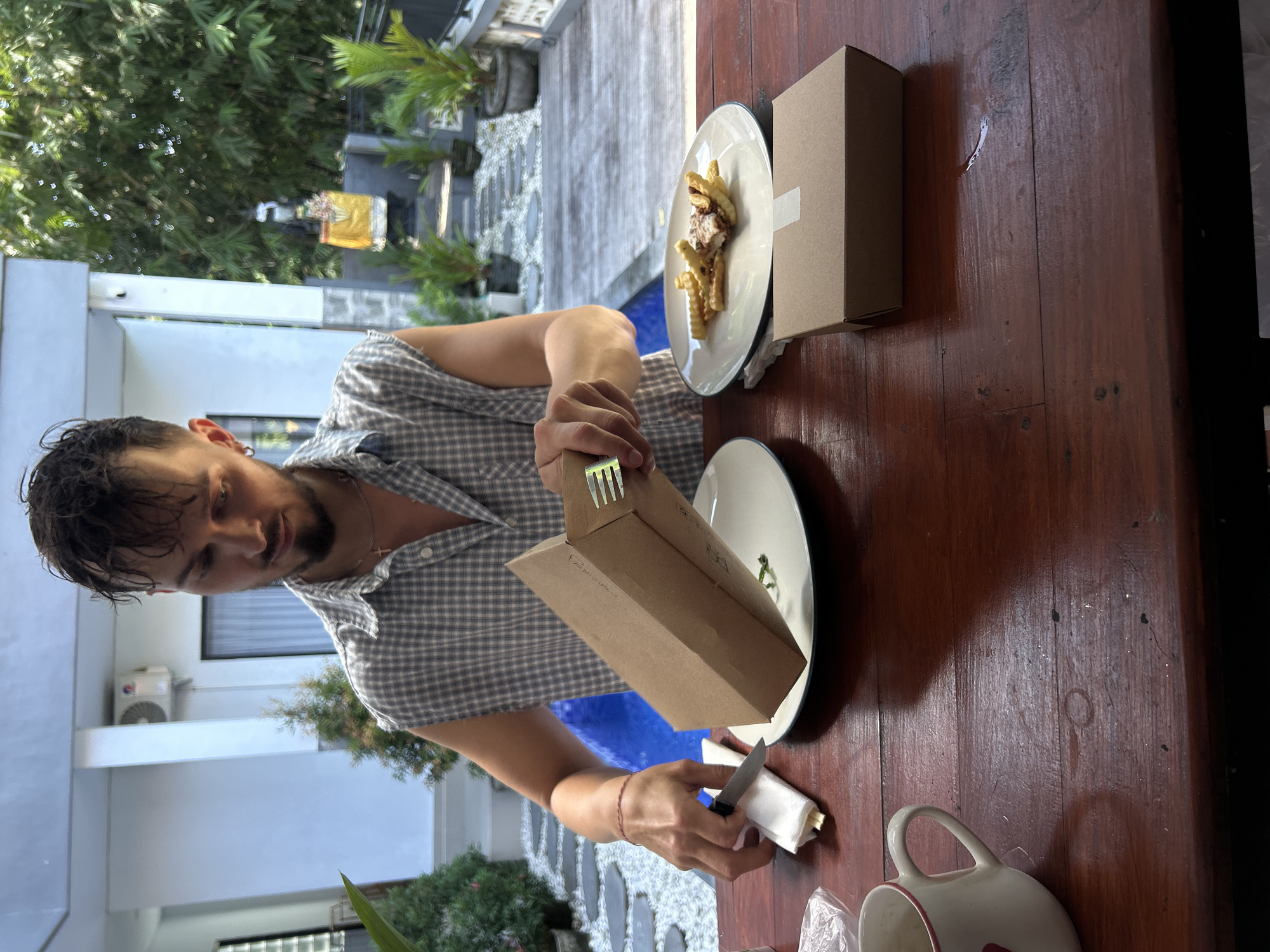 A man is shown from the chest up, sitting at a dark wooden table outdoors, in front of a swimming pool. He is wearing a grey plaid, sleeveless shirt and appears to be unpacking food from a cardboard box. In front of him, on a white plate, are French fries and what appears to be a small piece of chicken. To his left, a white napkin is folded with a knife. Another cardboard box is placed on the table to his right, in front of a potted plant. The background shows lush green foliage and architectural elements of a building. The overall setting suggests a meal being enjoyed in a tropical outdoor environment.