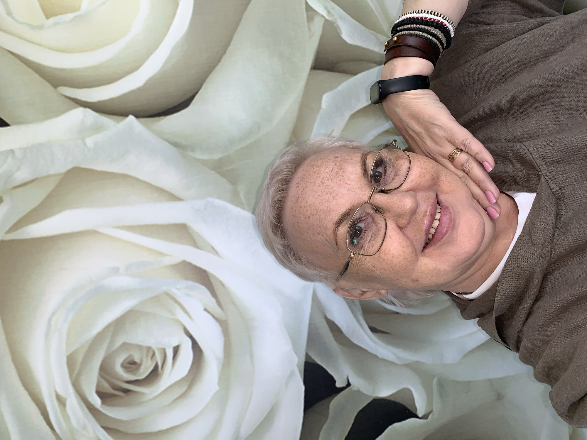 A middle-aged woman with short grey hair and glasses smiles at the camera. She is wearing a brown shirt and several bracelets on her left wrist, as well as a fitness tracker. Her left hand rests on her cheek, where she wears a gold ring. The background features a large, pale rose floral pattern. There is no indication of the location in the city of Chaikovsky, Russia.