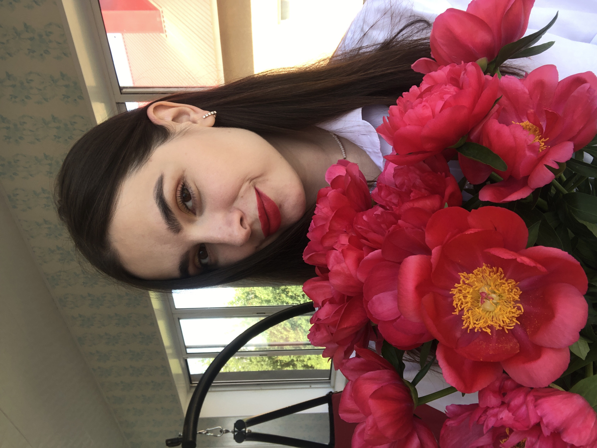 In a medium shot, a young woman with long, dark hair is holding a bouquet of vibrant red peonies. She is wearing a white collared shirt and a delicate pearl necklace. Her makeup includes bright red lipstick and subtle eyeshadow. She is positioned in front of a window with a view of trees and buildings, suggesting an outdoor or balcony setting. The background includes a patterned wallpaper and a hanging swing chair. The image appears to be taken in Noviy, Russia, possibly on a balcony or in a room with a view of the surrounding environment.