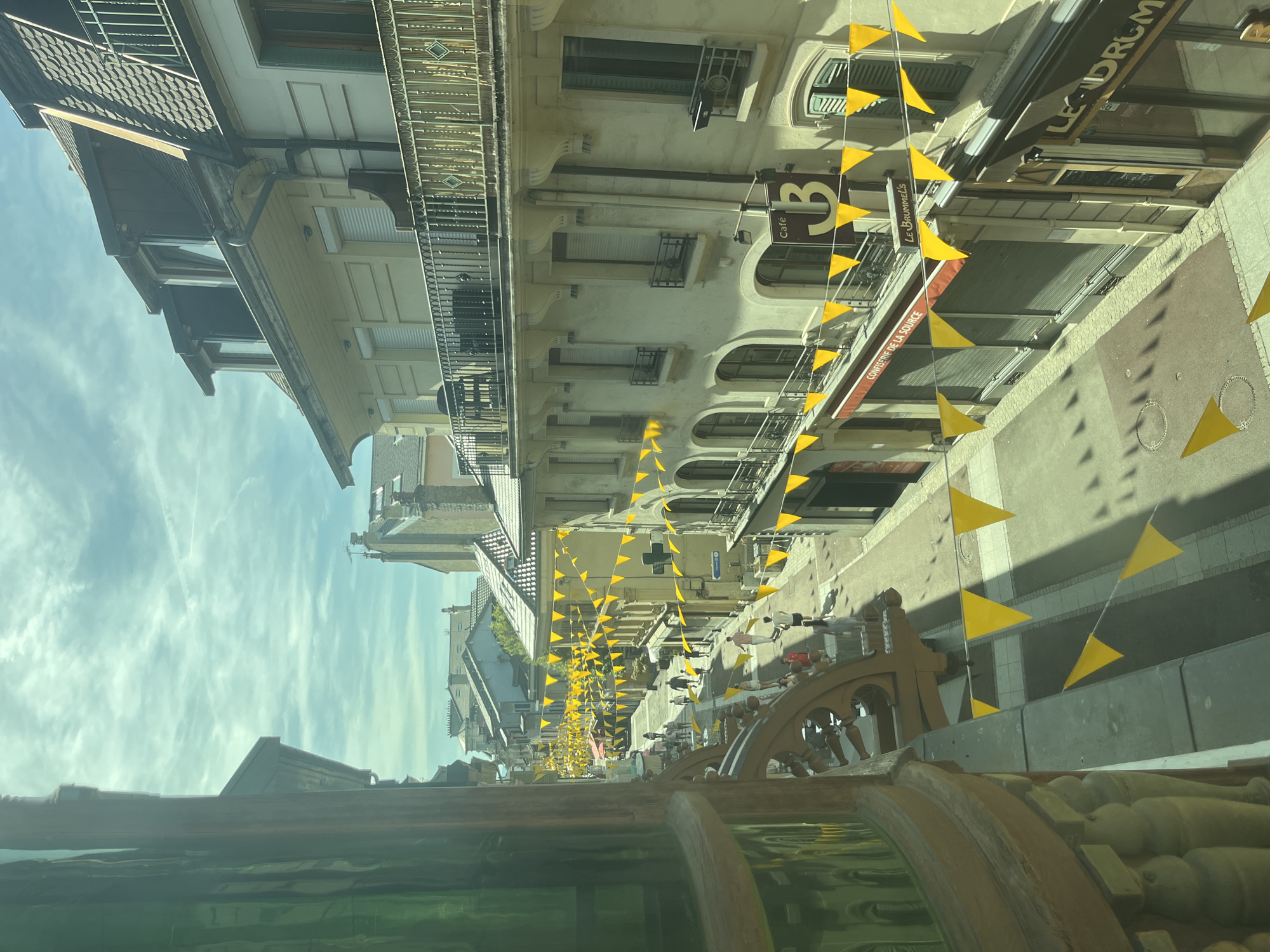 This is a medium-shot, eye-level, outdoor photograph taken on a sunny day in Évian-les-Bains, France. The image shows a street lined with buildings and decorated with yellow triangular flags strung across it.
In the foreground, on the left, a portion of a decorative architectural element with intricate carvings and green glass is visible. The street itself is paved and shows distinct shadows cast by the buildings and the flags. Several people are walking along the street, some singly and some in small groups. They are dressed in casual summer clothing.
The buildings on either side of the street are multi-story structures with awnings, balconies, and shuttered windows. Some of the buildings house shops and cafes, as indicated by signage such as "Confiserie de la Source," "Le Brummels," and "Cafe 3." The sky above is partly cloudy with streaks of white clouds. The overall atmosphere suggests a lively, festive day in a European town.