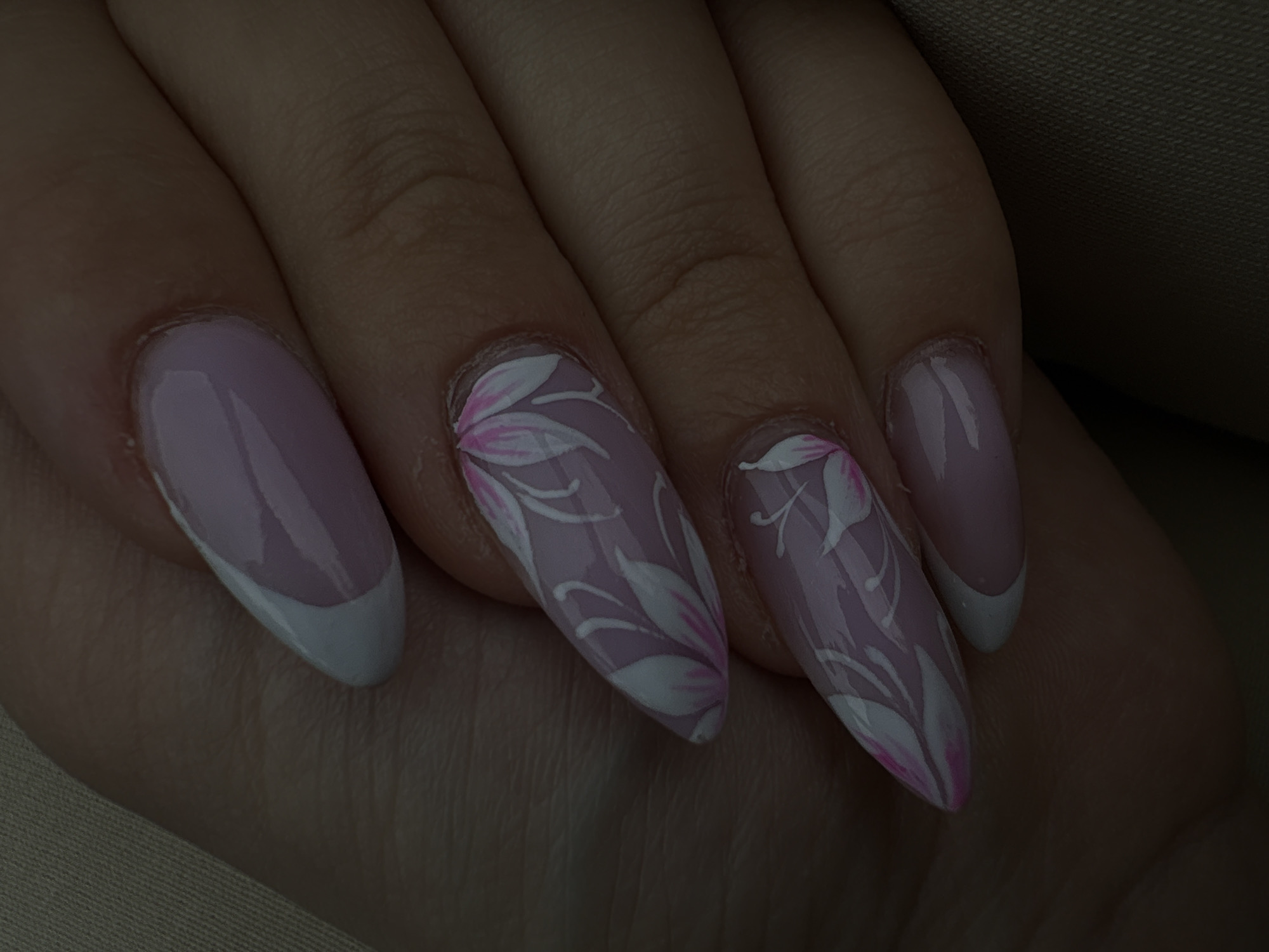 The image shows a close-up of a hand with four manicured nails. The nails have a light pink base. Two nails feature a classic French manicure with a white tip. The other two nails have a floral design in white and pink paint. The background is a soft, neutral color fabric. There is no indication that the image was taken in Plovdiv, Bulgaria.