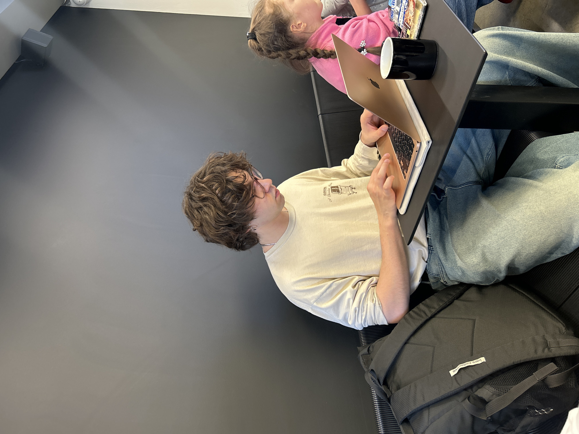 This indoor photograph, taken in Izhevsk, Russia, features a young person and a child in what appears to be a modern cafe or study area. The main subject is a young person with curly brown hair, wearing glasses, a light-colored long-sleeve shirt with a faint "A Chocolate Bar" graphic, and blue jeans. They are seated, actively using a gold-colored MacBook laptop that rests on a dark gray table. A silver chain is visible around their neck.
To their side, partially visible behind the laptop, is a young child with braided hair, wearing a pink long-sleeve top. A black mug and a small package of snacks are also on the table near the child. The background consists of a plain, dark gray wall, suggesting a minimalist and contemporary setting. A black backpack with a white tag that reads "iliars collective" is placed on the floor next to the main subject. The lighting is bright and even, indicative of daytime. The young person appears focused on their laptop activity.
