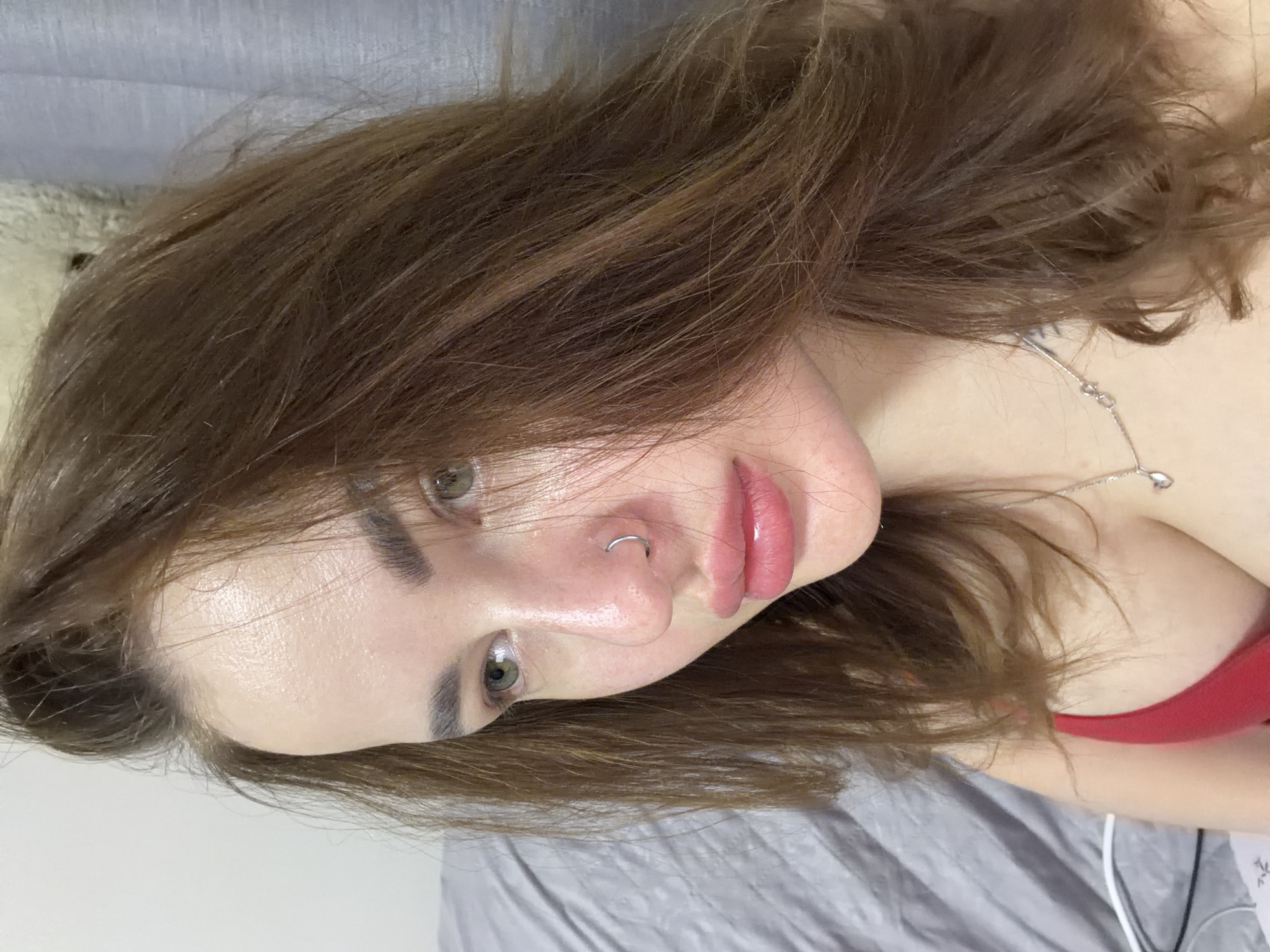 A young woman with brown hair and green eyes is lying on a gray bed. She has a silver nose ring and a silver necklace. She is wearing a red top. The background includes a gray wall and a glimpse of a white stuffed animal. The image does not provide information to confirm the location as Krasnodar, Russia.