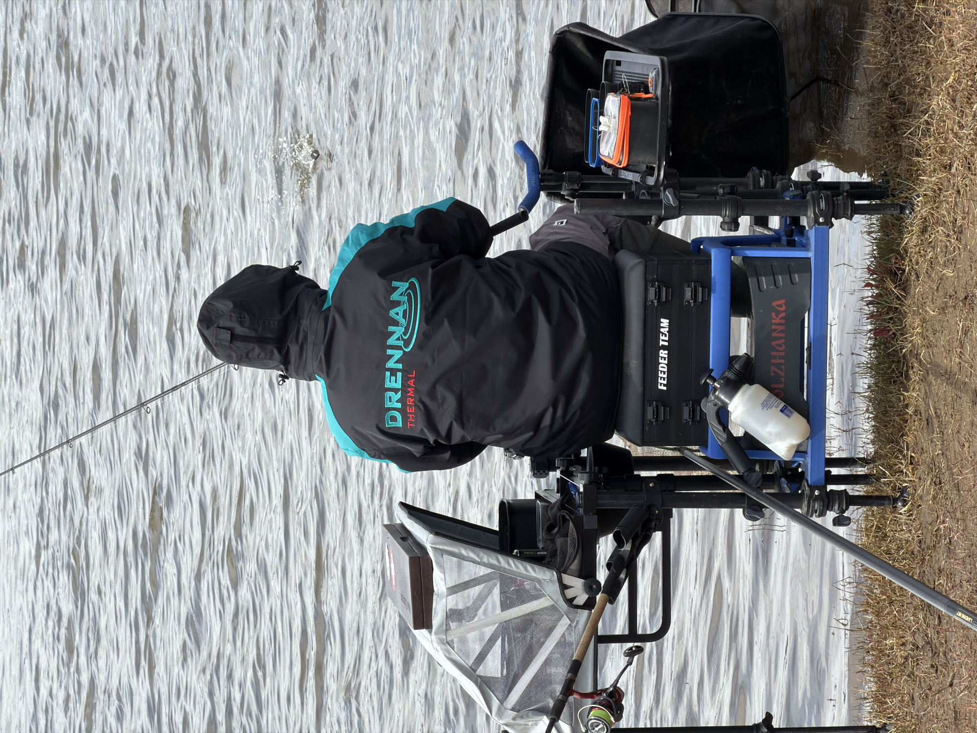 A person wearing a black jacket with "Drennan Thermal" printed on the back is fishing. The person is sitting on a blue and black fishing chair equipped with various fishing accessories and equipment. The fishing chair is placed on the edge of a body of water with visible ripples on the surface. To the right of the chair is dry, grassy land. The scene is likely located in Nikolo-Beryozovka, Russia, although this cannot be definitively confirmed from the image.