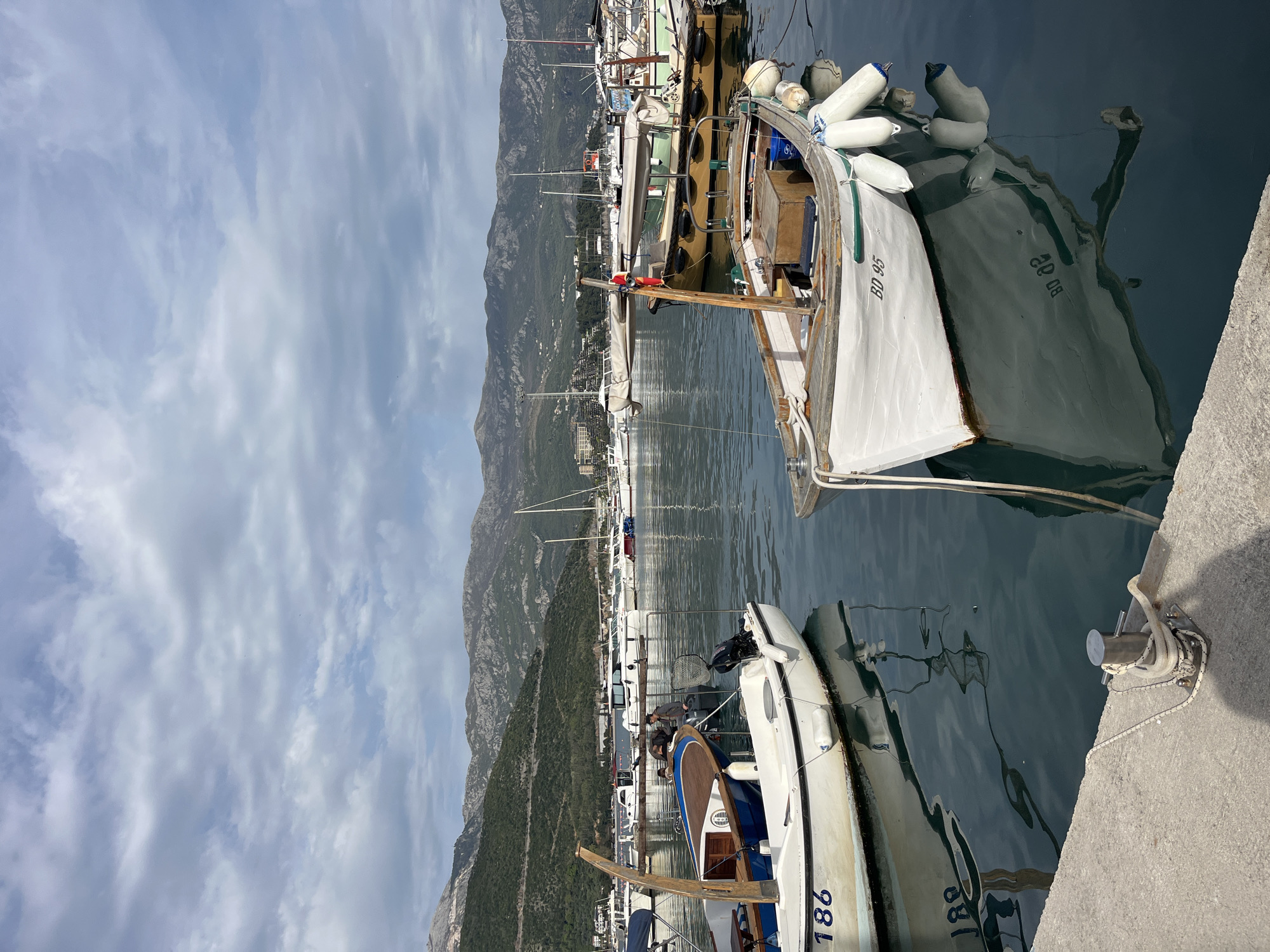 The image shows a marina in Budva, Montenegro. Several boats are docked along a concrete pier. In the foreground, there are two white boats. One boat has the registration number "BD 95" and is equipped with white fenders. The second boat displays the number "186". In the background, there is a view of mountains and a town with buildings and boats. The sky is cloudy. The water in the marina is calm.