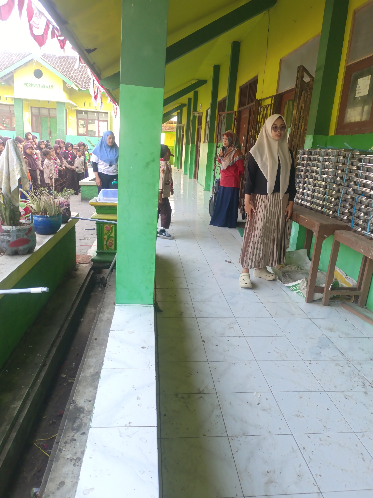 The image depicts an outdoor scene on a tiled walkway in front of a building with a green and yellow facade. In the foreground, a person wearing a cream-colored hijab, a black long-sleeved top, and a pleated skirt stands facing forward. To their left, another person in a red shirt and blue skirt, also wearing a hijab, holds a microphone. Behind them, a group of children in school uniforms are visible, some wearing hijabs. A building with "PERPUSTAKAAN" (library) written on it is visible in the background, suggesting the location is likely a school in Sarimulyo, Indonesia. A wooden bench with stacks of metal containers is positioned to the right of the walkway. The walkway tiles are dirty, with visible debris. The environment appears to be outdoors, possibly a school hallway or courtyard, with green painted pillars and a yellow wall.