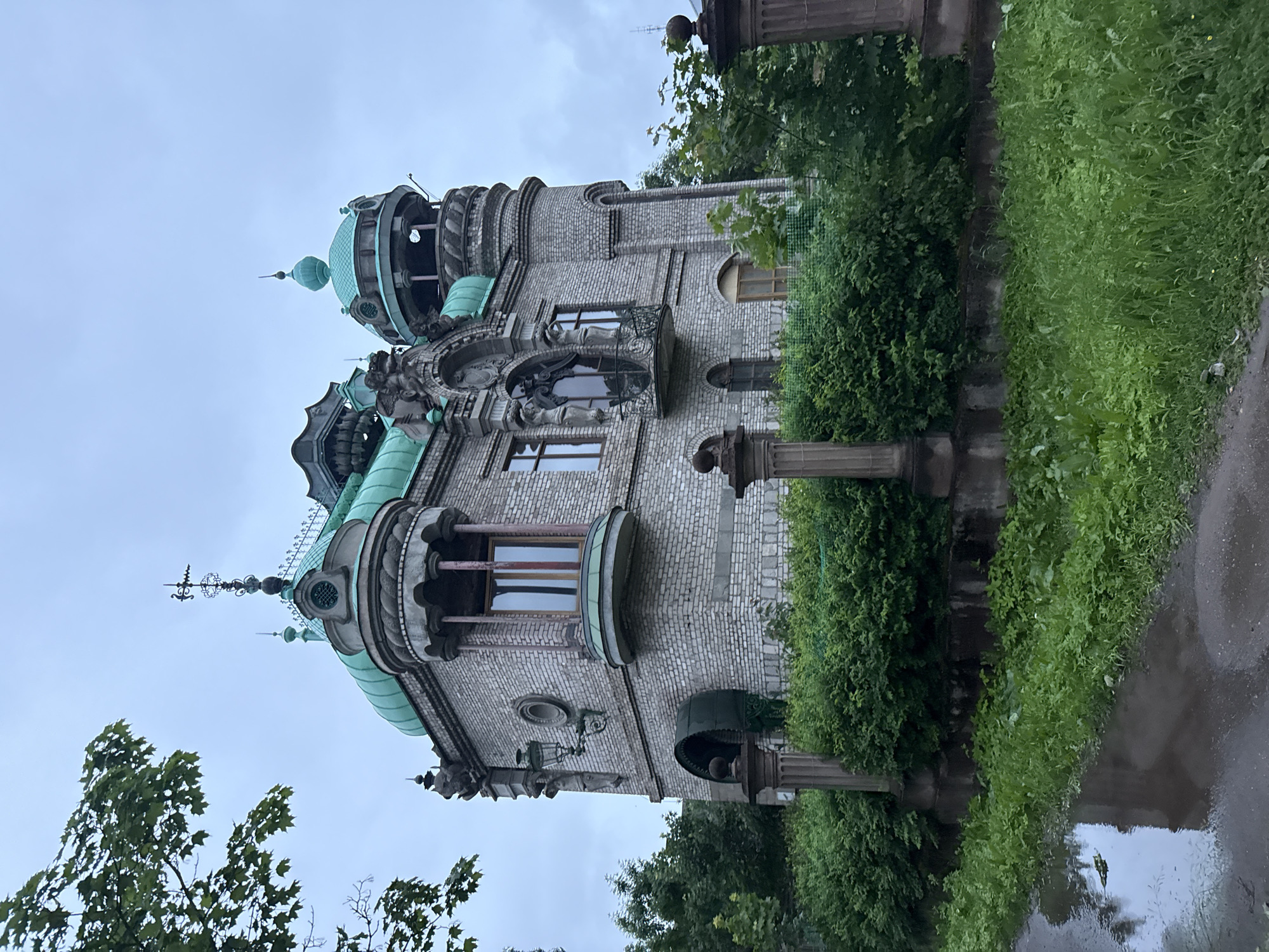 This image shows an exterior view of the Terentyev House, located in the city of Valday, Russia. The building is a unique architectural structure with a grey brick facade and multiple green domes and turrets. It features ornate details, including statues, decorative window surrounds, and a wrought-iron balcony. The house is surrounded by dense green bushes and foliage. In the foreground, a wet, muddy path with puddles suggests recent rain. The sky is overcast and grey. There are no people or animals visible in the image. The scene appears to be captured on a cloudy day.