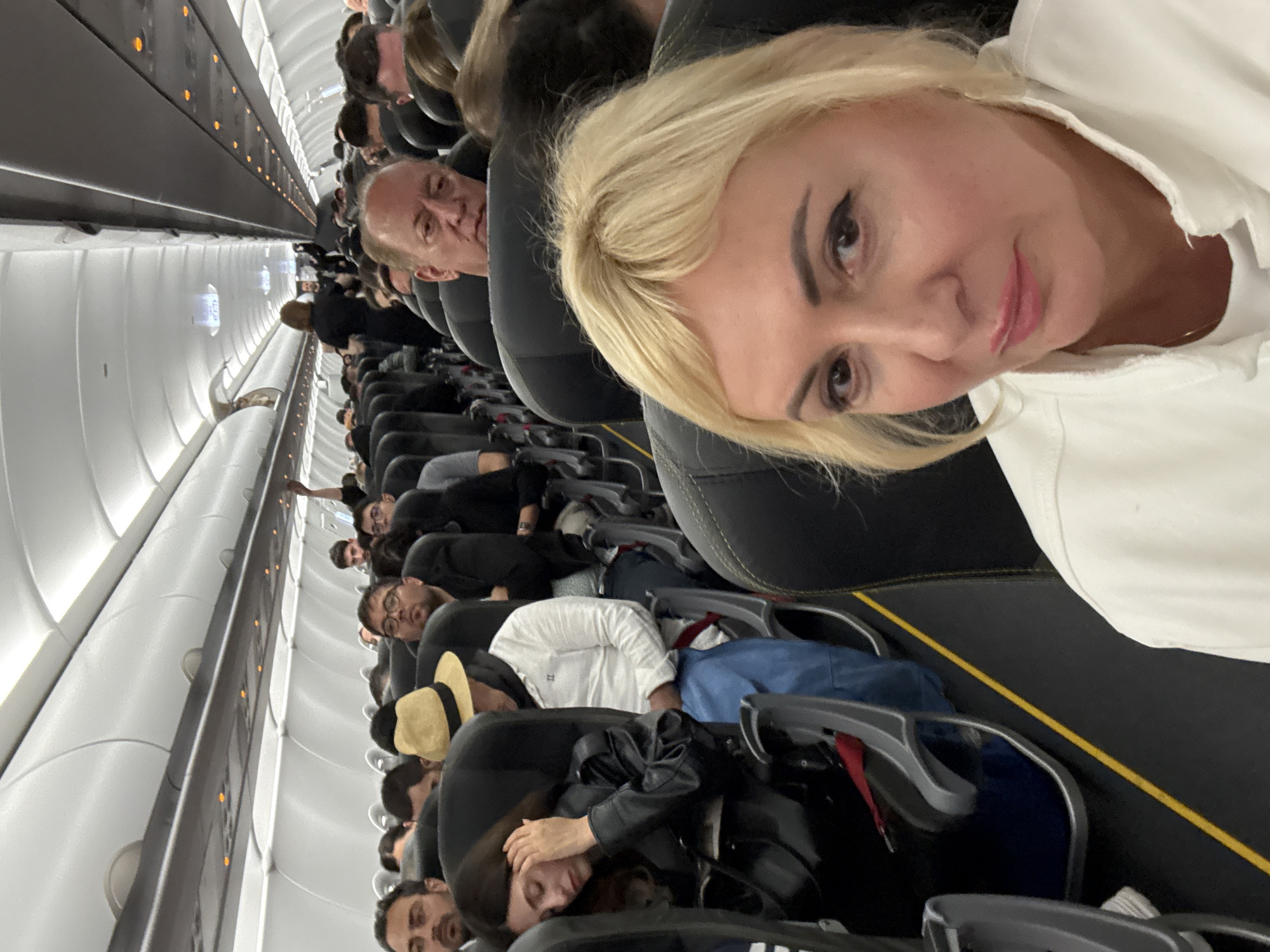 The image shows the interior of a commercial airplane cabin. The aircraft appears to be fully occupied with passengers seated in rows of black seats, most wearing dark clothing. A woman with blonde hair is prominently positioned in the foreground, possibly taking a selfie. A man can be seen looking towards the camera behind her. The cabin's overhead compartment is visible on the left side of the frame. The location is Unknown, Unknown.