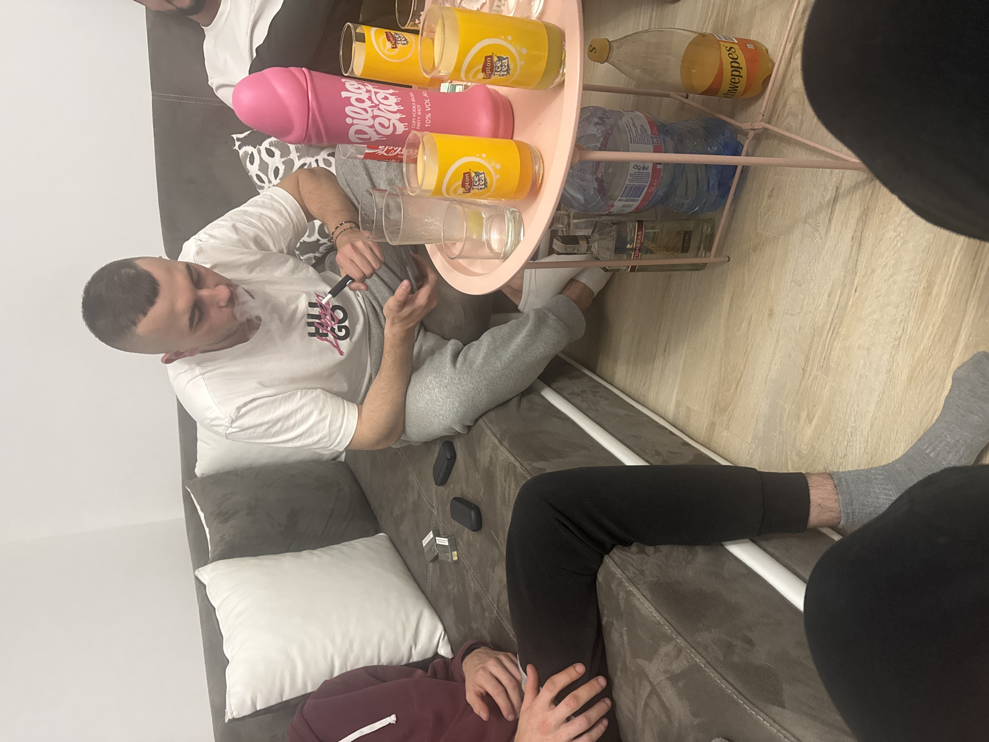 The image depicts an indoor scene featuring a group of people. A fair-skinned man with short hair is sitting on a brown couch, holding a black object resembling a vaping device, with smoke visible around his mouth. He is wearing a white t-shirt with graphic text. On a nearby pink side table, there are multiple Lipton Ice Tea glasses, a pink novelty "Dildo Shot" bottle, a Schweppes bottle, water bottles, and what appears to be a bottle of Tumbras. A second person is partially visible, wearing black pants and a grey sock. Another individual, wearing a red hoodie, is partially visible near the bottom edge of the image. Cigarettes and a mobile phone can be seen on the couch. The setting appears to be a living room, with neutral-colored walls and flooring. The image does not contain sufficient information to determine the location as Graf Ignatievo, Bulgaria.