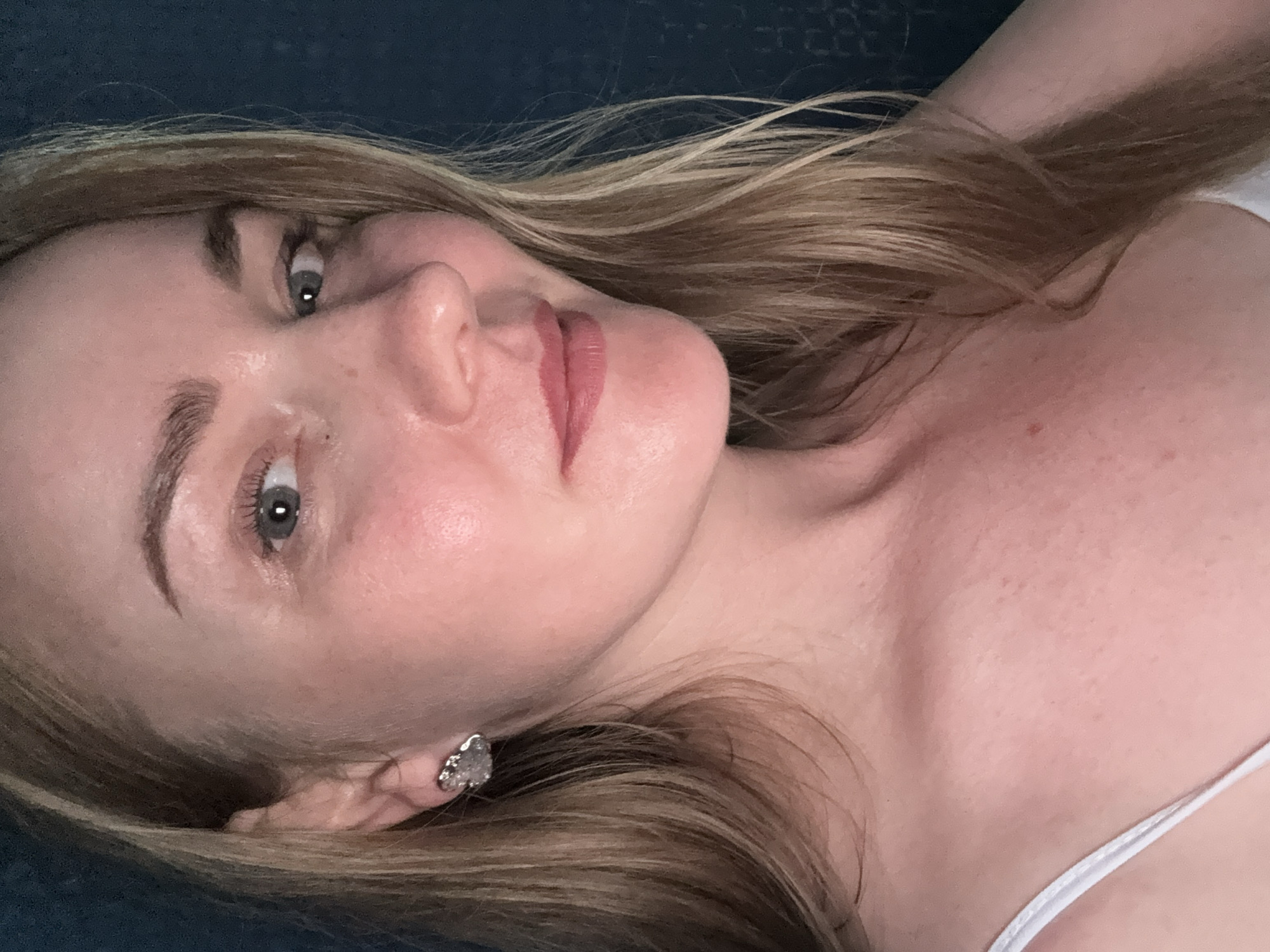 This is a close-up, waist-up, indoor, selfie-style photograph of a fair-skinned, blonde woman, possibly in her late 30s or early 40s. She is looking towards the right side of the frame with her head tilted slightly. Her hair is long, blonde, and appears to be slightly wavy, falling over her shoulders. She is wearing a white tank top. A single, silver, heart-shaped earring is visible on her left ear. Her makeup includes light foundation, possibly blush on her cheeks, defined eyebrows, and a muted pink lipstick. Her skin shows some texture and subtle blemishes. The background is a dark, possibly blue, textured surface, suggesting an indoor setting. There is no indication of the city of Chaikovsky, Russia, being present in the image, and no specific geographic markers are visible.