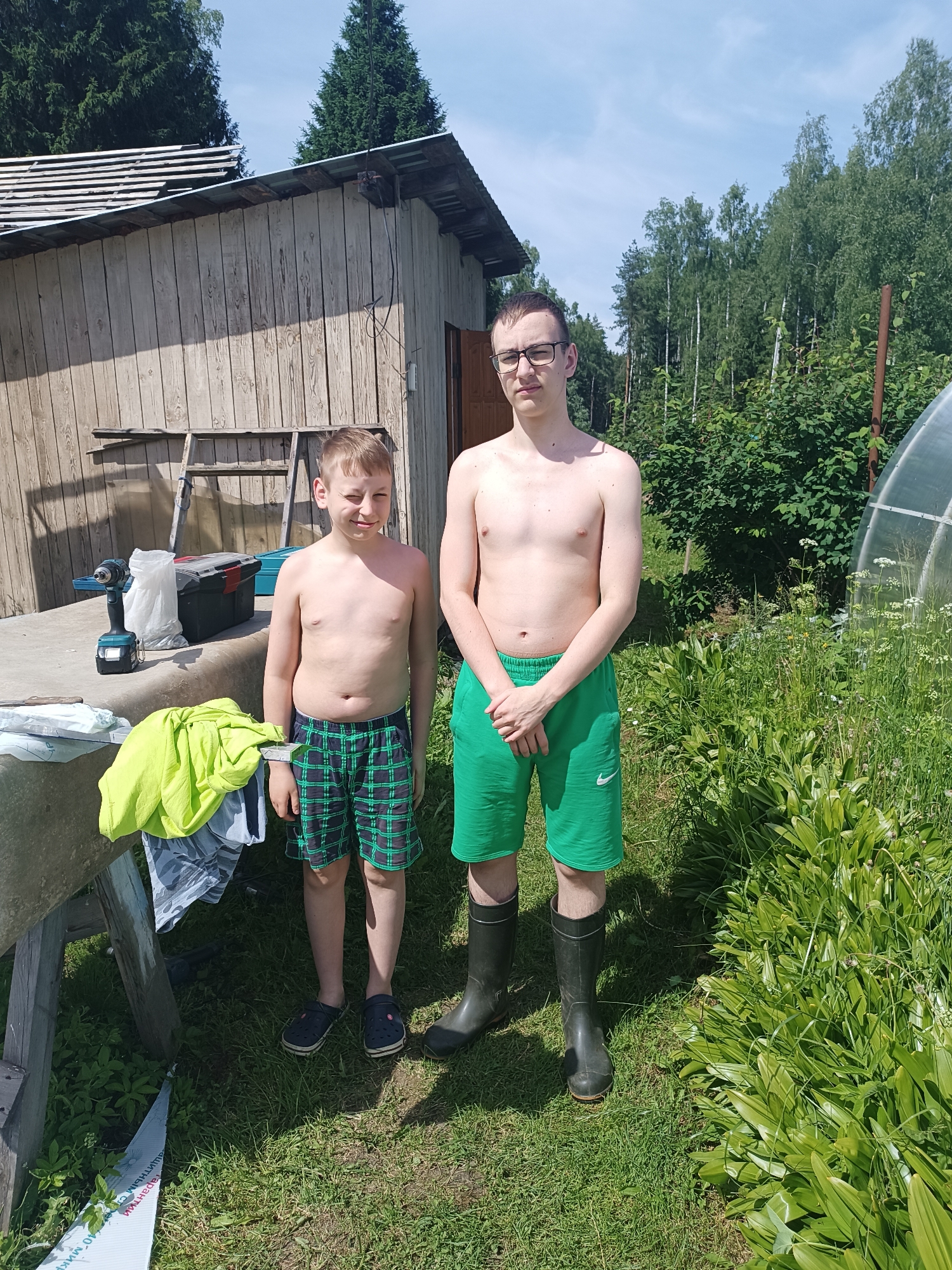 The image captures an outdoor scene in Новые Омутищи, Russia, under a clear blue sky with direct sunlight, casting distinct shadows. Two males are standing on a grassy area, appearing to pose for the camera.
The individual on the left is a male, estimated to be a pre-teen, with short, light-colored hair. He is shirtless and wearing green and black plaid shorts with dark blue slip-on shoes. He stands slightly left of the image center, facing the camera with his left eye partially closed and a slight upward turn of his lips.
The individual on the right is a male, estimated to be a young adult, with short, dark hair and black-framed glasses. He is shirtless and wearing plain green athletic shorts featuring a white Nike logo on the left thigh, along with dark green rubber boots. He stands to the right of the younger male, facing the camera with a neutral expression, his hands clasped in front of his waist.
In the background, behind the younger male, stands a light-colored wooden shed with vertical plank siding. The shed's roof appears to have exposed or unfinished structural elements on its left side. A wooden ladder leans against the shed wall. To the right, behind the older male, a translucent, arched greenhouse structure is partially visible. A dense treeline, comprising both coniferous and deciduous trees, forms the distant background.
In the mid-ground, to the left of the younger male, a rough, light-colored workbench or table is present. On its surface, a blue and black Makita cordless drill, a white plastic bag, a black toolbox, and a blue container are arranged, alongside a pile of clothing including a bright lime green garment. On the ground near the workbench leg, a piece of white material with Russian text is visible. A brown, vertical metal pole is situated in the mid-ground, right of the younger male. The ground beneath the individuals is covered with green grass and various plants, some displaying small white flowers.