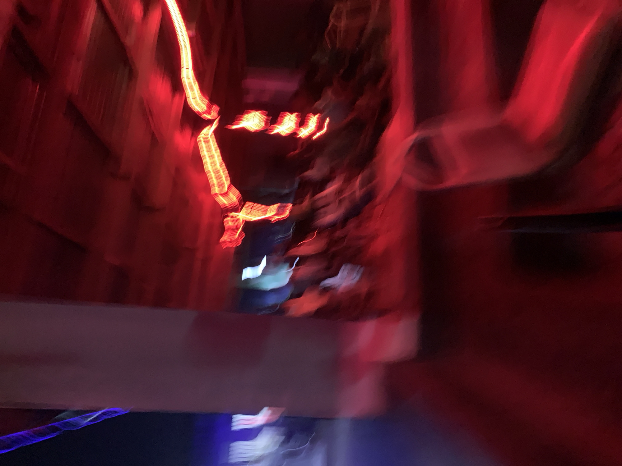 The image shows a blurred and distorted view of a street at night, likely taken while in motion. Predominantly red lighting illuminates the scene, with what appears to be neon signs and building facades. There is a horizontal structure, possibly a barrier or railing, with red and white colors near the lower portion of the image. Blue and white lights can be seen below this structure. The scene lacks clarity, and specific details regarding people, objects, or actions are difficult to discern. The location of New Stadium, Nigeria, is not visually verifiable within the frame.