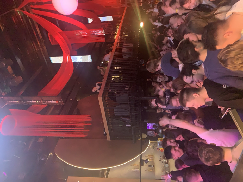 The image captures a crowded indoor scene at Gipsy Nightclub, located on Bolotnaya Embankment in Moscow, Russia. The venue is multi-level, with a densely packed main floor and an elevated mezzanine/balcony overlooking it. Red fabric drapes and long vertical red tassels adorn the ceiling, extending downwards. A large, circular, bronze-toned wall panel with an illuminated neon outline is prominent on the left, adjacent to a dimly lit bar counter featuring various bottles. Approximately 50 to 70 individuals, predominantly young adults of mixed gender, occupy the space. On the main floor, people are standing in close proximity, many with arms raised, suggesting active participation in dancing or celebration. On the mezzanine, fewer individuals are visible, observing the activity below or socializing near the dark metal railing. The lighting is dim, utilizing spotlights and ambient colored lights, primarily casting red and purple hues across the scene. Large speakers are mounted high on the walls, integrated into the structure. The overall environment appears lively and energetic.