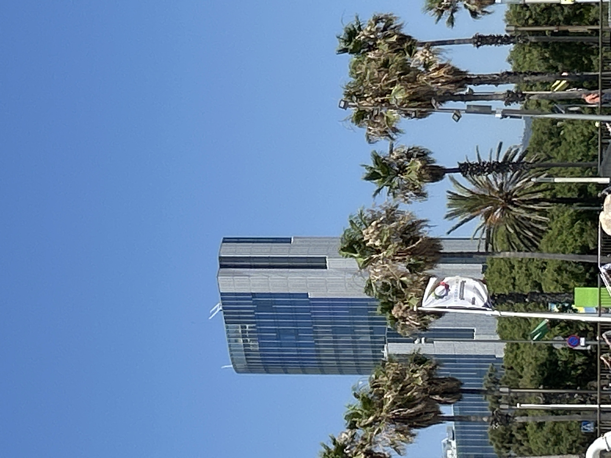 The image captures a sunny day in Barcelona, Spain, with a clear blue sky overhead. In the background, a tall, modern glass and steel skyscraper dominates the skyline. In the foreground, a row of lush palm trees sways gently. A white flag with a colorful circular logo and text is planted in the ground, fluttering in the breeze. The scene suggests a vibrant urban environment with a touch of tropical flair, possibly a waterfront area or a park.