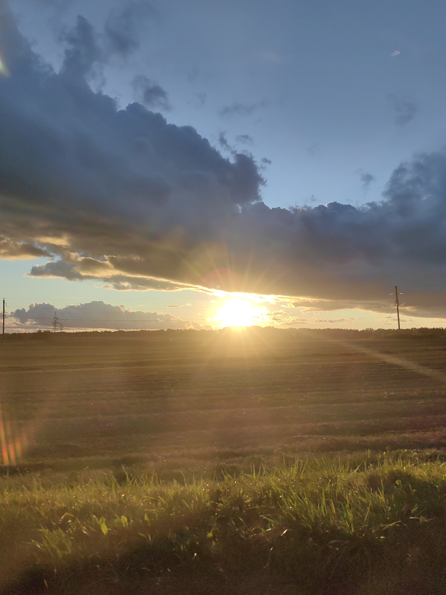 The image captures a sunset over a rural landscape in the vicinity of Фаніпальскі сельскі Савет, Belarus. The sky is dramatic, with dark, heavy clouds on the left side contrasting with bright, illuminated clouds and the intense glow of the setting sun on the right. The sun's rays are bursting through a gap in the clouds, casting a warm, golden light across a vast, flat field. The field appears to be agricultural, possibly recently harvested, with visible rows or textures suggesting cultivation. In the foreground, a strip of lush green grass lines the bottom edge of the frame, catching the last light of the day. Two utility poles with power lines are visible in the distance, one on the left and another on the right, indicating the presence of infrastructure in the otherwise natural setting. The overall scene is one of natural beauty and tranquility, with a hint of the approaching evening.