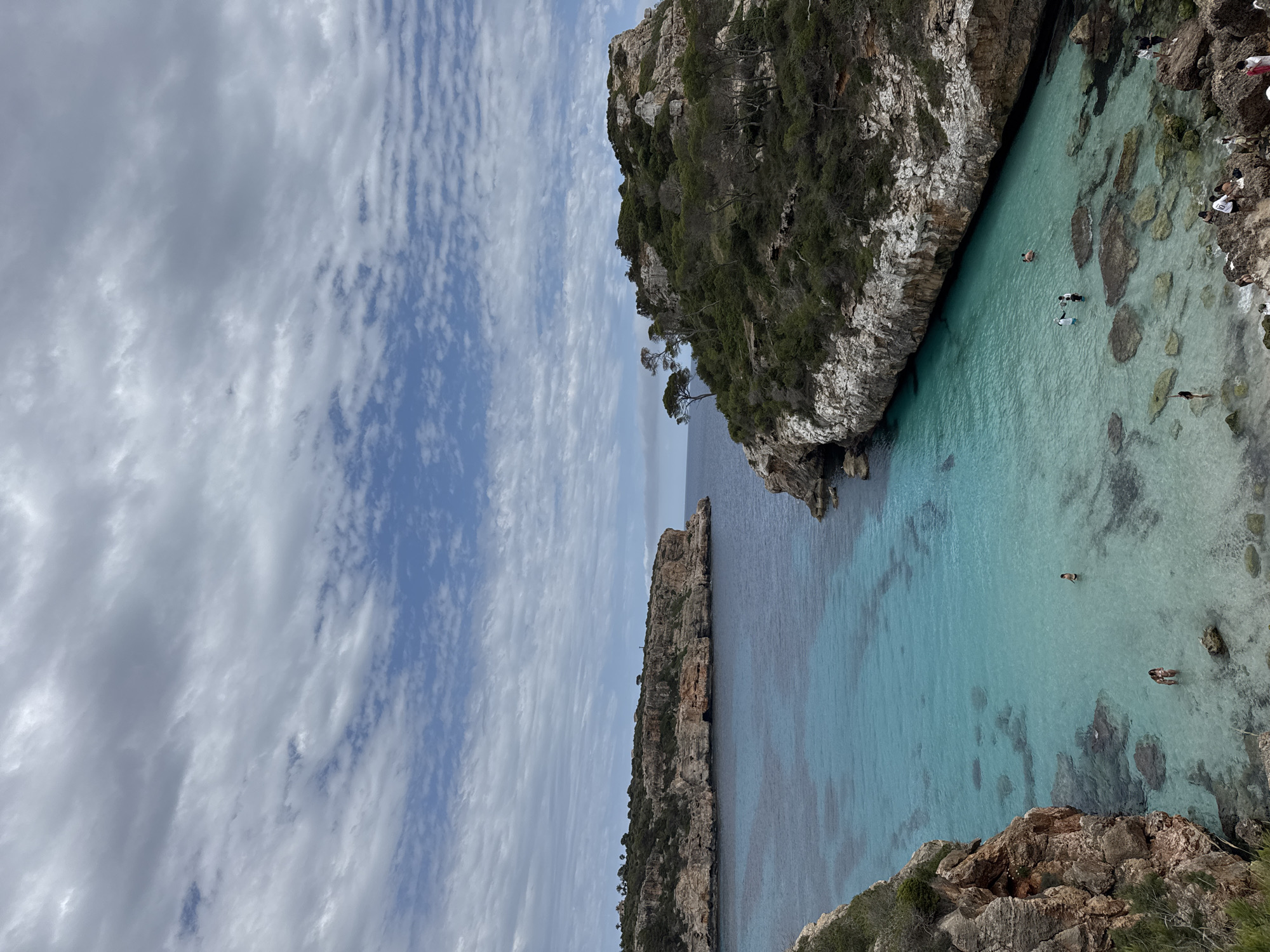The image presents a view of Cala des Moro in Santanyí, Spain. The landscape is dominated by steep, rocky cliffs covered with vegetation on either side, enclosing a narrow bay with turquoise water. A few individuals are visible swimming or wading in the clear, shallow water near the rocky edges. The sky is partially cloudy, with a mix of white and grey formations. The rocks display a textured surface with varying shades of brown and grey. The water reflects the sky.