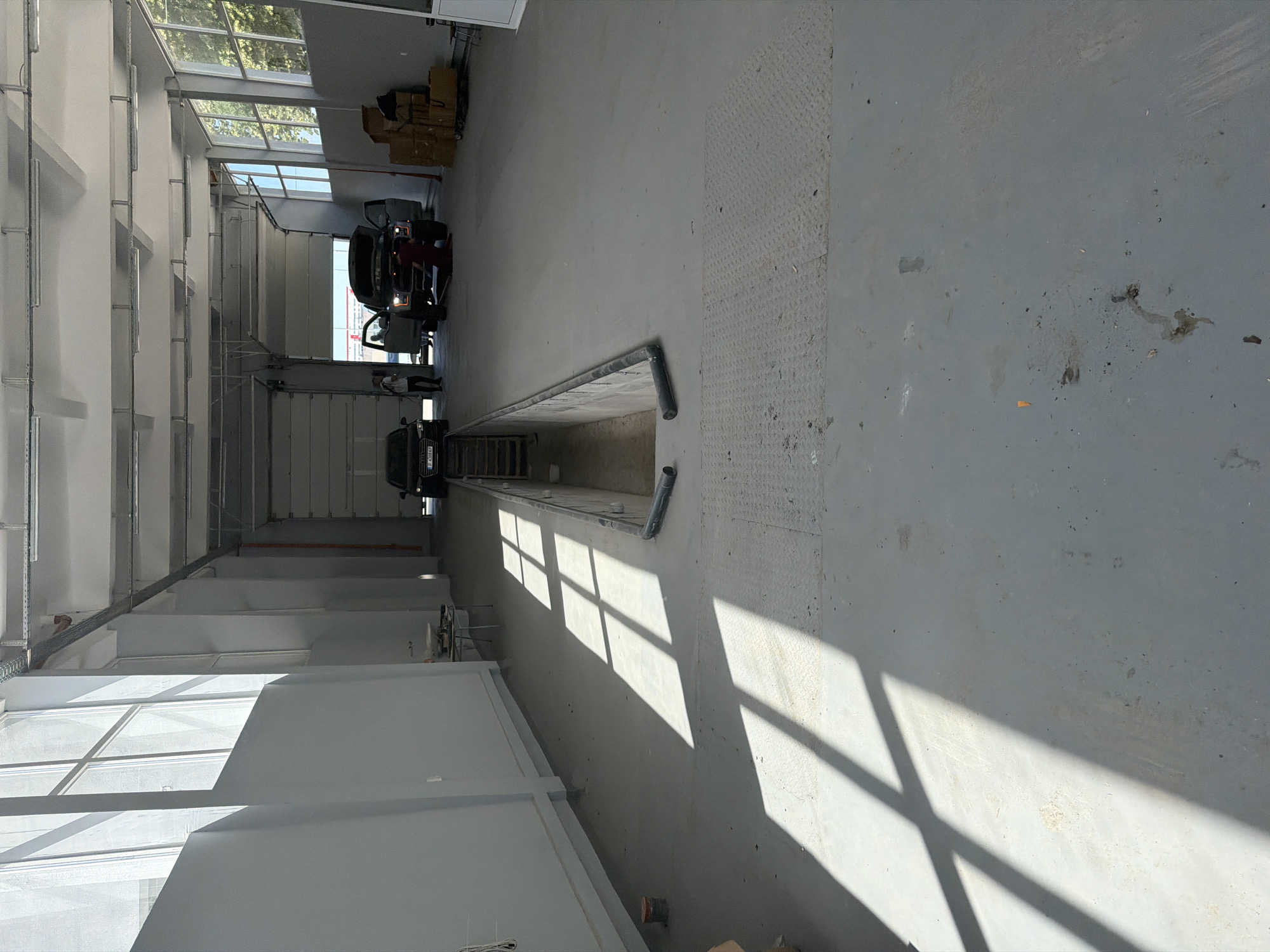 An interior view of a commercial garage or workshop in Ruse, Bulgaria. The space is well-lit with natural light streaming from large windows along the left wall and overhead fluorescent lighting. The floor is a light gray concrete, showing some stains and debris. In the foreground, shadows from the windows cast a grid pattern across the floor.
A recessed pit, likely for vehicle maintenance, is visible in the middle ground, with a metal staircase leading down into it. Two vehicles are present: a dark-colored sedan parked to the left of the pit and a pickup truck, possibly a Ford Raptor, with its hood open, positioned over the pit on the right. Several people are near the pickup truck, appearing to be engaged in work or conversation.
To the right of the pickup truck, stacked cardboard boxes suggest storage or recent deliveries. The walls are painted a light gray, with some exposed conduit and pipes on the ceiling. A large, rolled-up garage door is visible in the background, indicating the entrance to the facility. The overall impression is of an active, functional automotive service area.