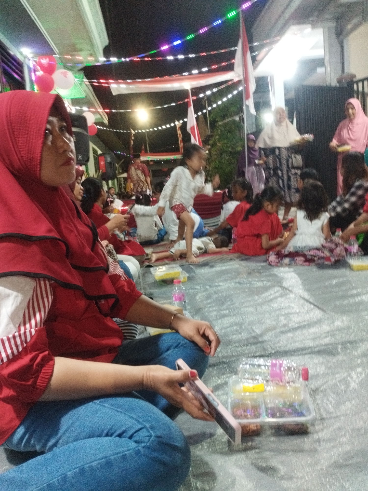 The image depicts a gathering of people, possibly a celebration or community event, taking place outdoors at night. The environment is decorated with colorful string lights and Indonesian flags, suggesting a festive occasion. The ground is covered with a silver-colored tarp, and people are seated or lying on it.
In the foreground, a woman wearing a red hijab and a red and white striped shirt is prominently featured. She is looking to her left, and her hands are resting on her lap, holding a pink smartphone. Next to her, a clear plastic container holds what appear to be small food items or snacks, with a pink and red bottle of water placed behind it.
In the mid-ground and background, several individuals are visible. A group of children are seated on the tarp, some in red and white clothing. One child in a white dress is sitting on a patterned rug. In the further background, adults are also present. One woman in a light-colored hijab is holding a plate, and another in a pink hijab is standing. The overall atmosphere appears to be relaxed and communal. While the specific location in Surabaya, Indonesia, cannot be determined from the image alone, the presence of Indonesian flags strongly indicates the country.