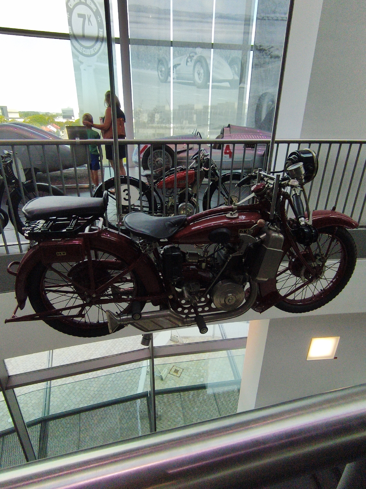 This is a photograph of a vintage motorcycle displayed at the Audi Forum in Ingolstadt, Germany. The motorcycle is dark red and is suspended by metal supports. It features chrome accents, a dark seat, spoked wheels, and a prominent headlight. In the background, visible through a glass partition, are other vehicles, including a classic race car. A woman and a young boy are standing near the glass, looking at the display. The Audi Forum is an automotive museum and visitor center located in Ingolstadt, Bavaria, Germany.
