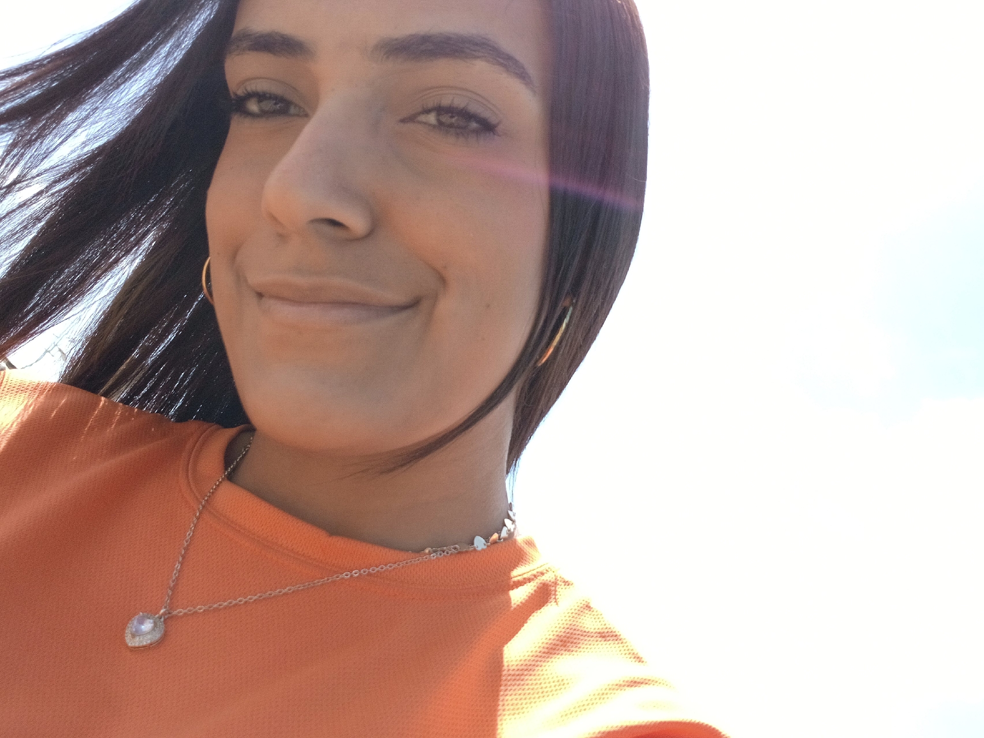 A woman is seen in a close-up shot. Her hair is dark and blowing in the wind, and she is wearing gold hoop earrings. She has on an orange shirt, with a silver necklace. The photo appears to be taken outdoors, with a bright sky. The location is not discernible from the image.