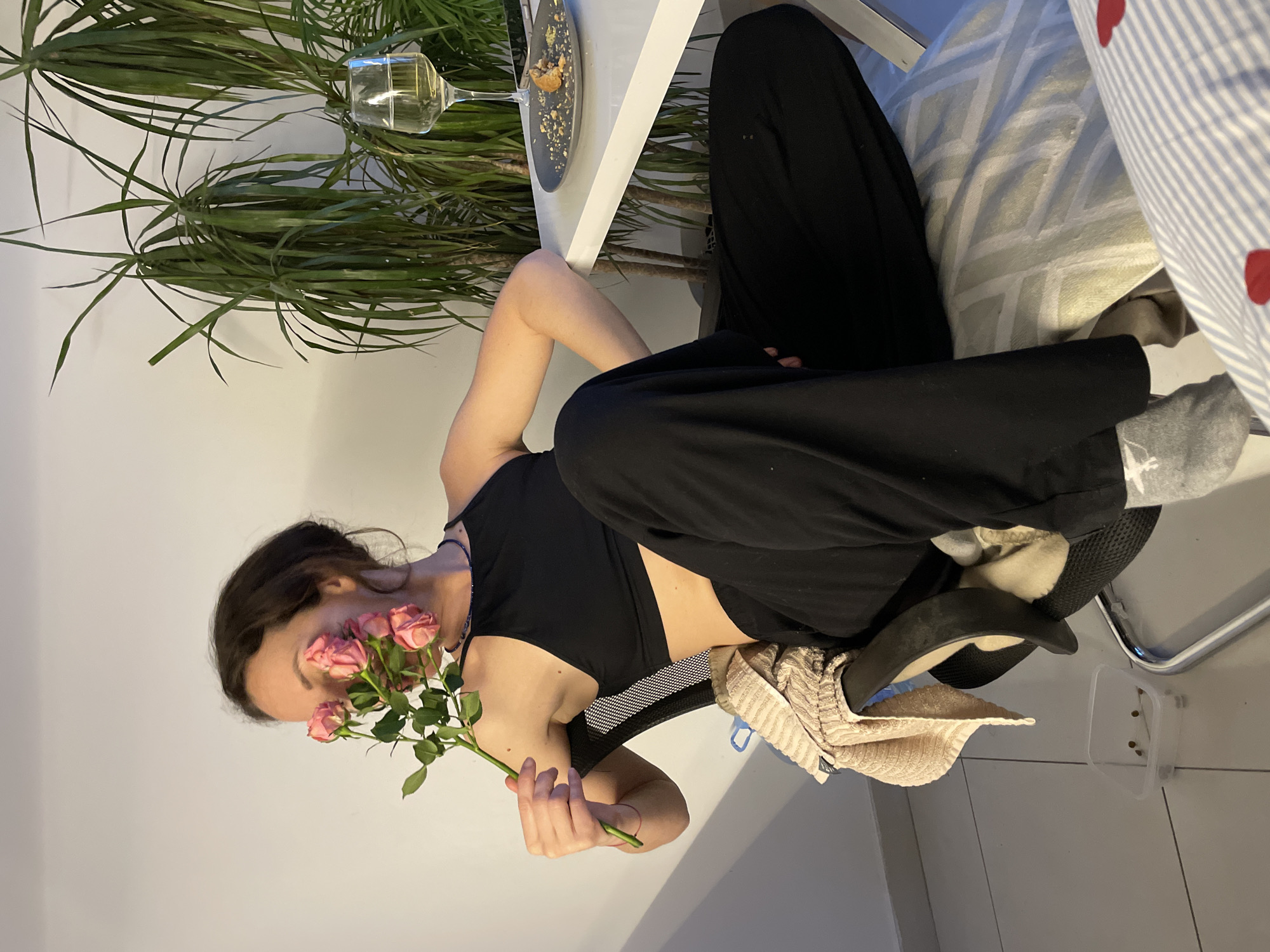 An indoor, horizontally oriented shot depicts a young woman seated, holding a small bouquet of pink roses to her face, partially obscuring it. She wears a black tank top and black wide-leg pants. Her dark hair is casually styled.
To her left (viewer's right, if oriented correctly), a white table holds a wine glass with a light-colored beverage and a dark plate with crumbs. Behind the table and the woman is a large potted plant with long, slender green leaves, possibly a Dracaena. The woman's right arm rests on the table.
To her right (viewer's left), patterned cushions or bedding are visible, including one with blue and white stripes and red heart patterns. Her feet are partially visible, wearing gray socks. A black sandal or slipper is on the floor near her feet, along with a light-colored, textured cloth draped over the chair. A small, clear plastic container with brown pellets, likely pet food, is also on the floor.
The lighting suggests an indoor setting, possibly evening, with artificial light illuminating the scene. The overall ambiance is relaxed and casual. The scene takes place in Boreti, Montenegro.